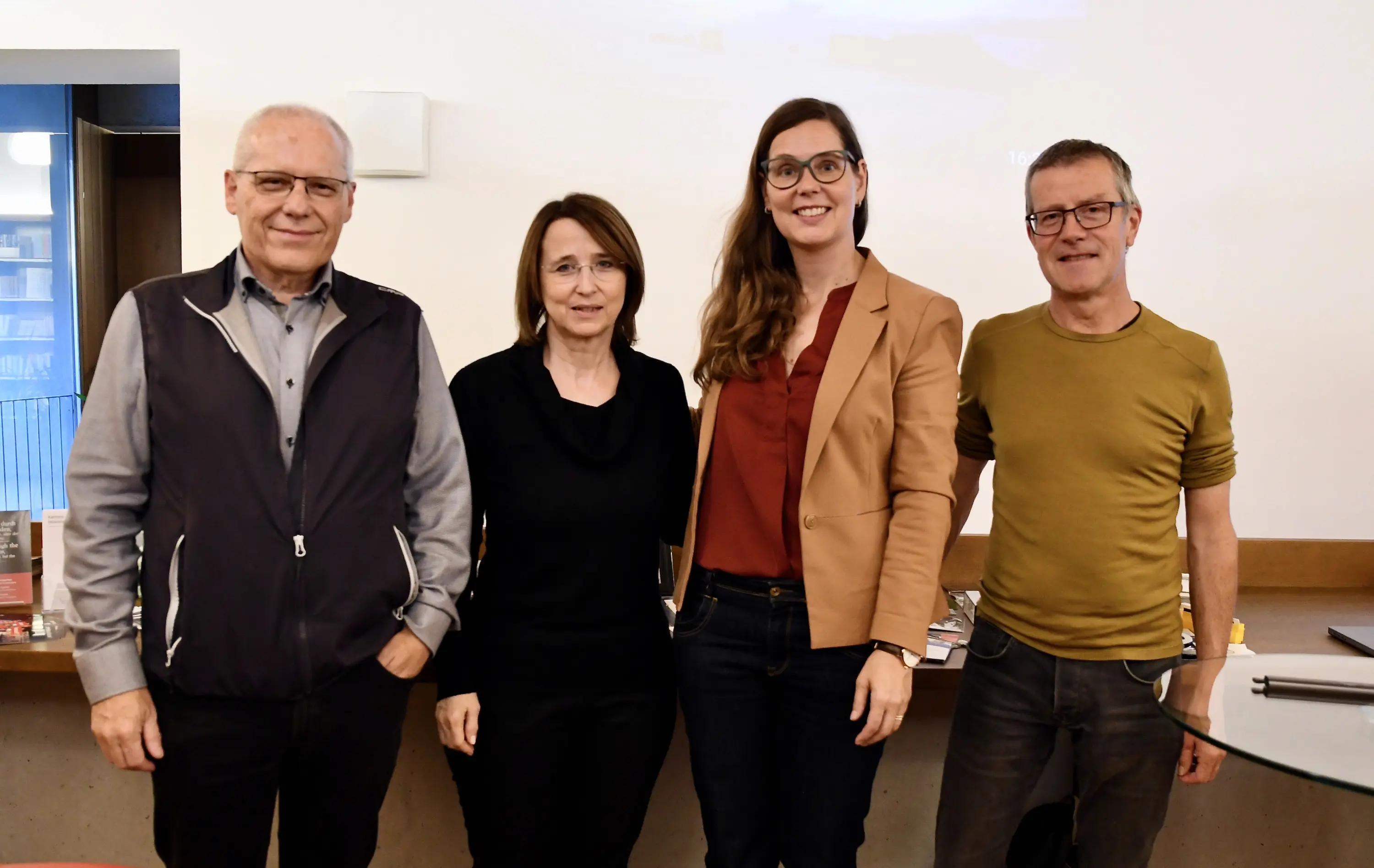 Quattro persone sorridenti si trovano in un ambiente interno, con un tavolo davanti. Due uomini e due donne, vestiti in modo casual. Sullo sfondo si intravedono alcuni oggetti e un progetto visivo.