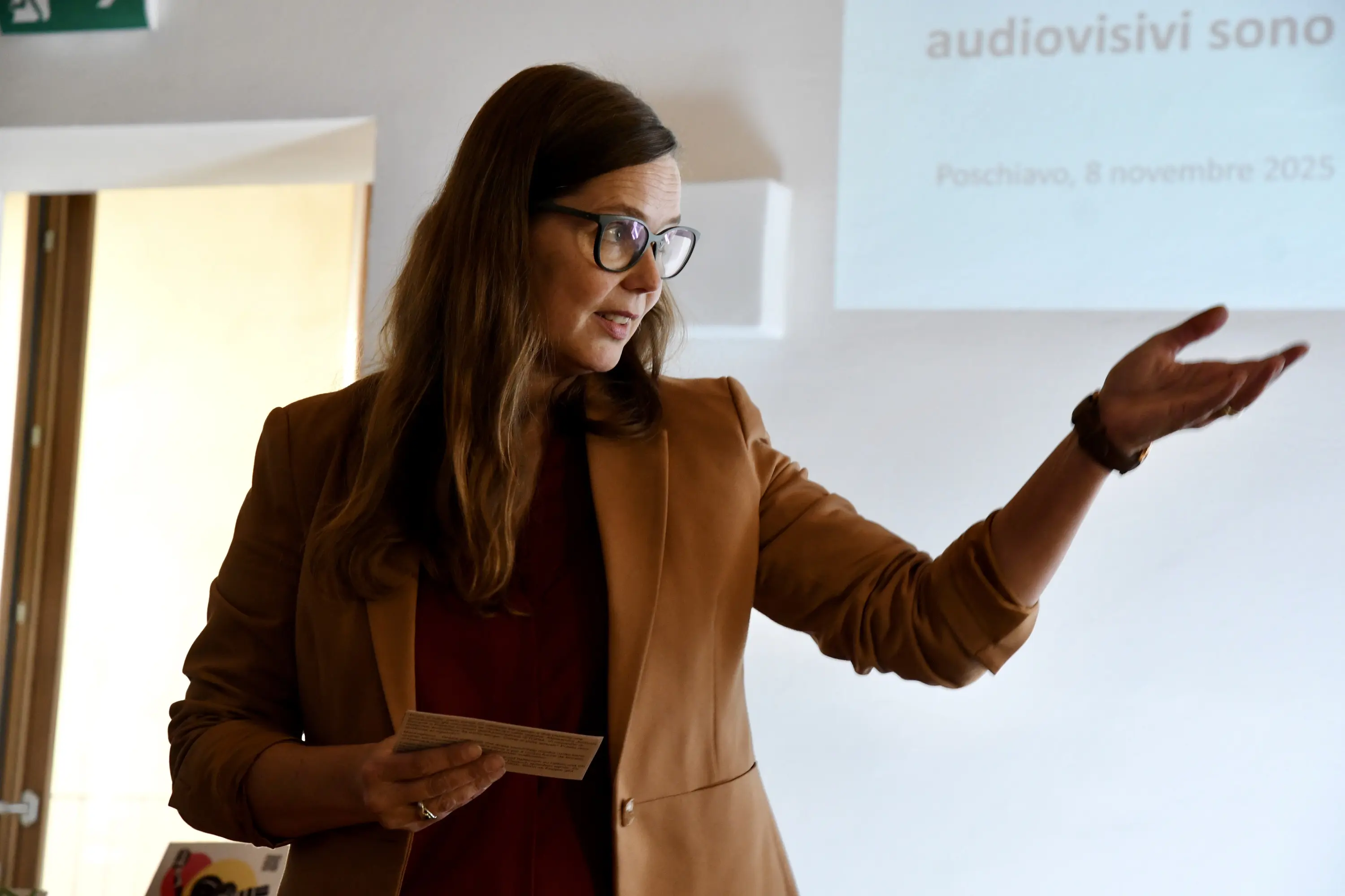 Una donna con occhiali e capelli lunghi, indossa un blazer marrone mentre parla e gesticola. Sullo sfondo si intravede un proiettore con testo visibile.