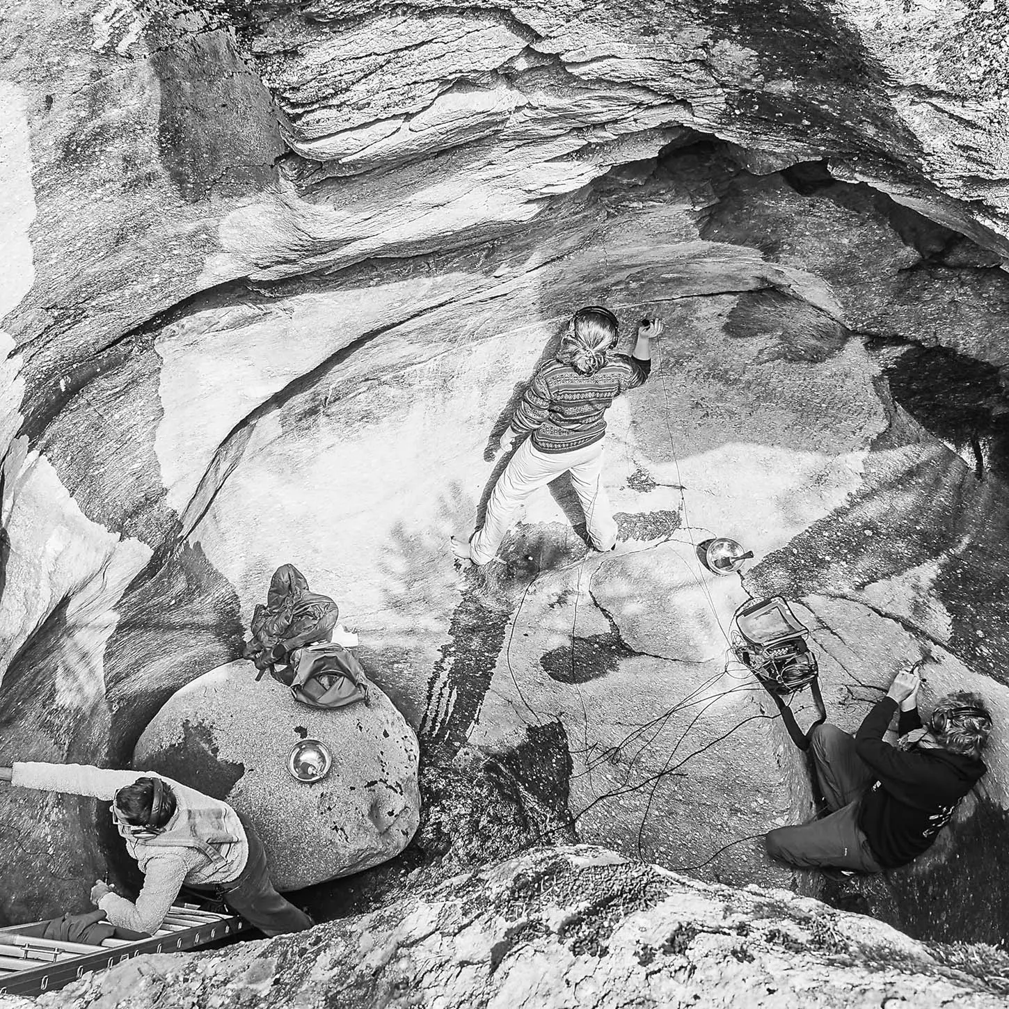 Persone impegnate in un'attività di arrampicata in una cavità rocciosa, con una di esse che scala la parete verticale. Altre due persone si trovano più in basso, una di fronte a una grande roccia e l'altra che sembra fornire