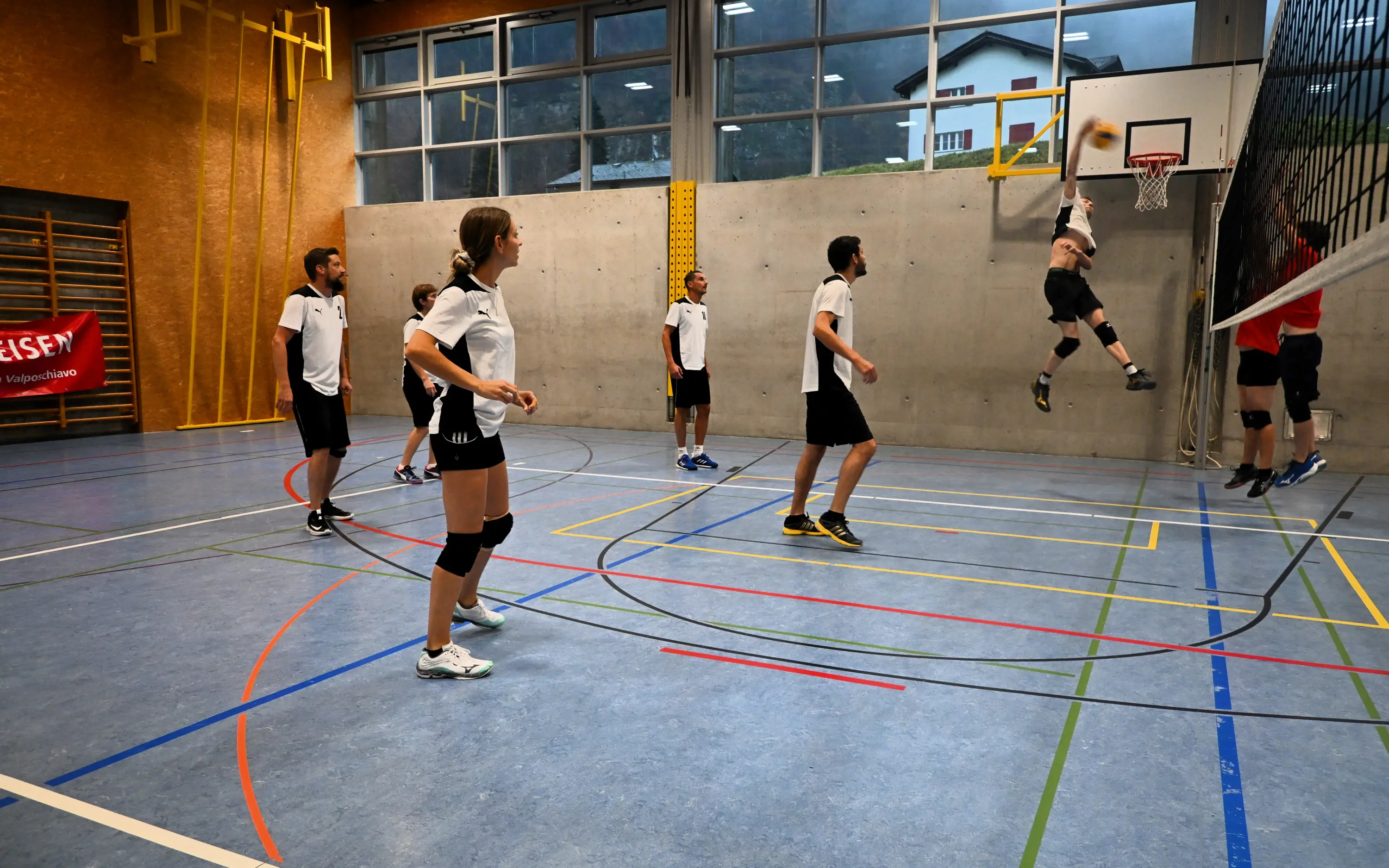 Giovani atleti che giocano a pallavolo in un campo al coperto. Un giocatore sta saltando per schiacciare la palla, mentre altri si preparano a riceverla. Ambiente luminoso con pareti in cemento e una rete.