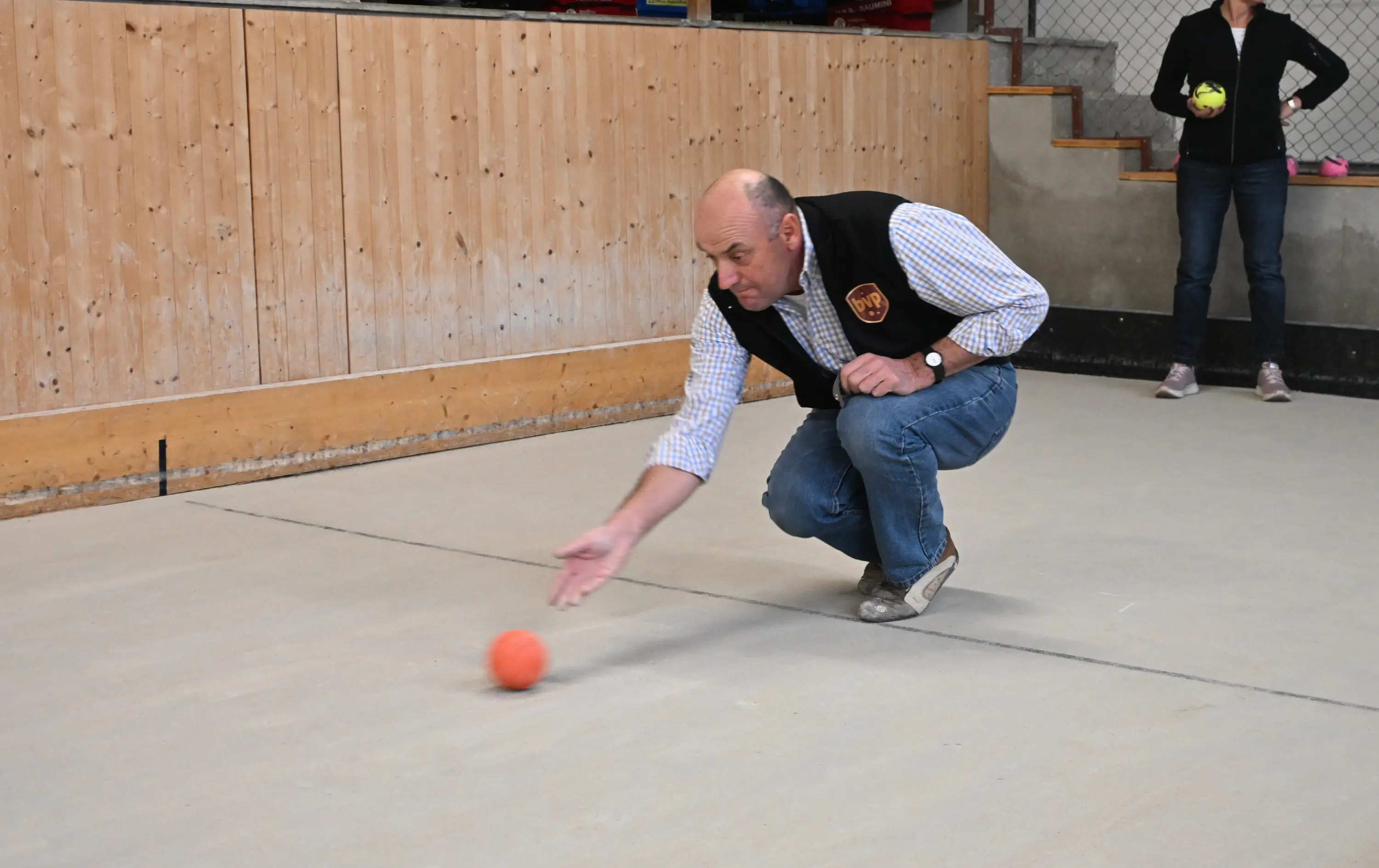 Un uomo si trova in ginocchio mentre lancia una pallina arancione su un campo di bocce. Indossa un gilet nero e una camicia a righe. Sullo sfondo, un'altra persona osserva, tenendo una pallina verde.