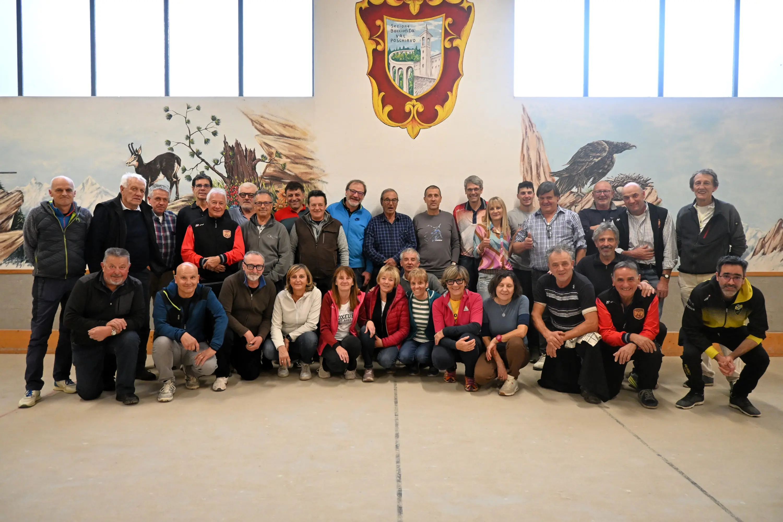 Gruppo di persone sorridenti in una sala con un murale sullo sfondo. Ci sono uomini e donne di diverse età, alcuni vestiti casualmente, in posa per la foto di gruppo.