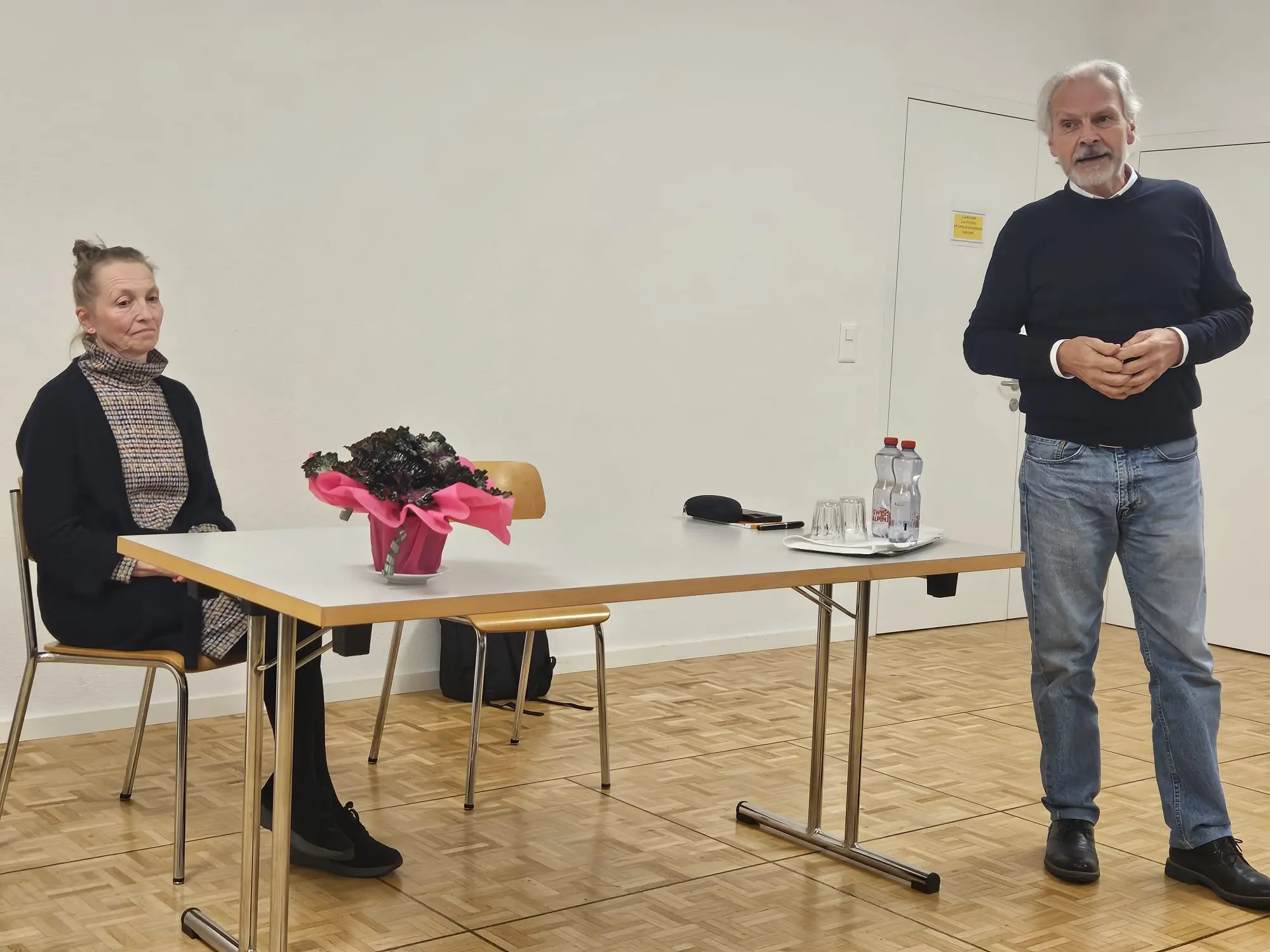 Due persone in una sala, una seduta e l'altra in piedi mentre parla. Sul tavolo c'è un vaso di fiori e delle bottiglie d'acqua.