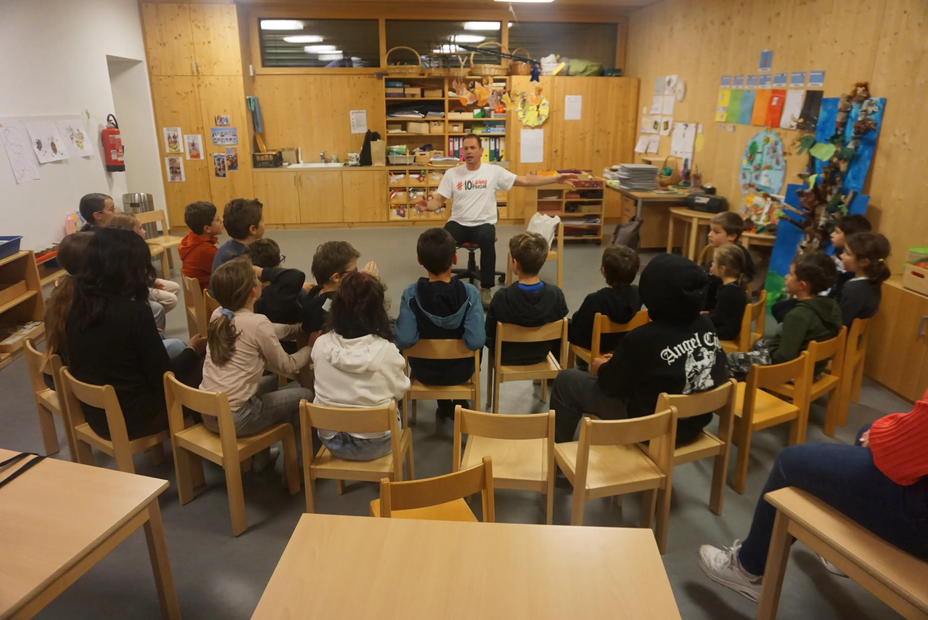 Un gruppo di bambini seduti in cerchio in una stanza accogliente, ascoltano un narratore che si trova di fronte a loro, seduto su una sedia. Gli adulti sono presenti ai margini del cerchio. Muri in legno e particolari decorazioni sulla