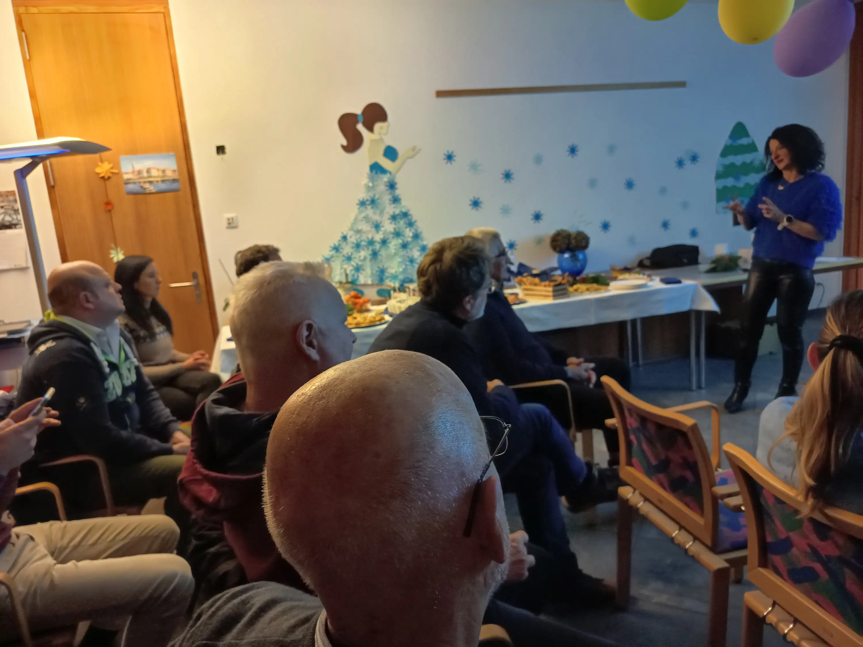 Un gruppo di persone sedute in una stanza durante un evento. Una donna in piedi parla davanti a loro, mentre una tavola imbandita con cibo si vede sullo sfondo. Decorazioni natalizie adornano le pareti.
