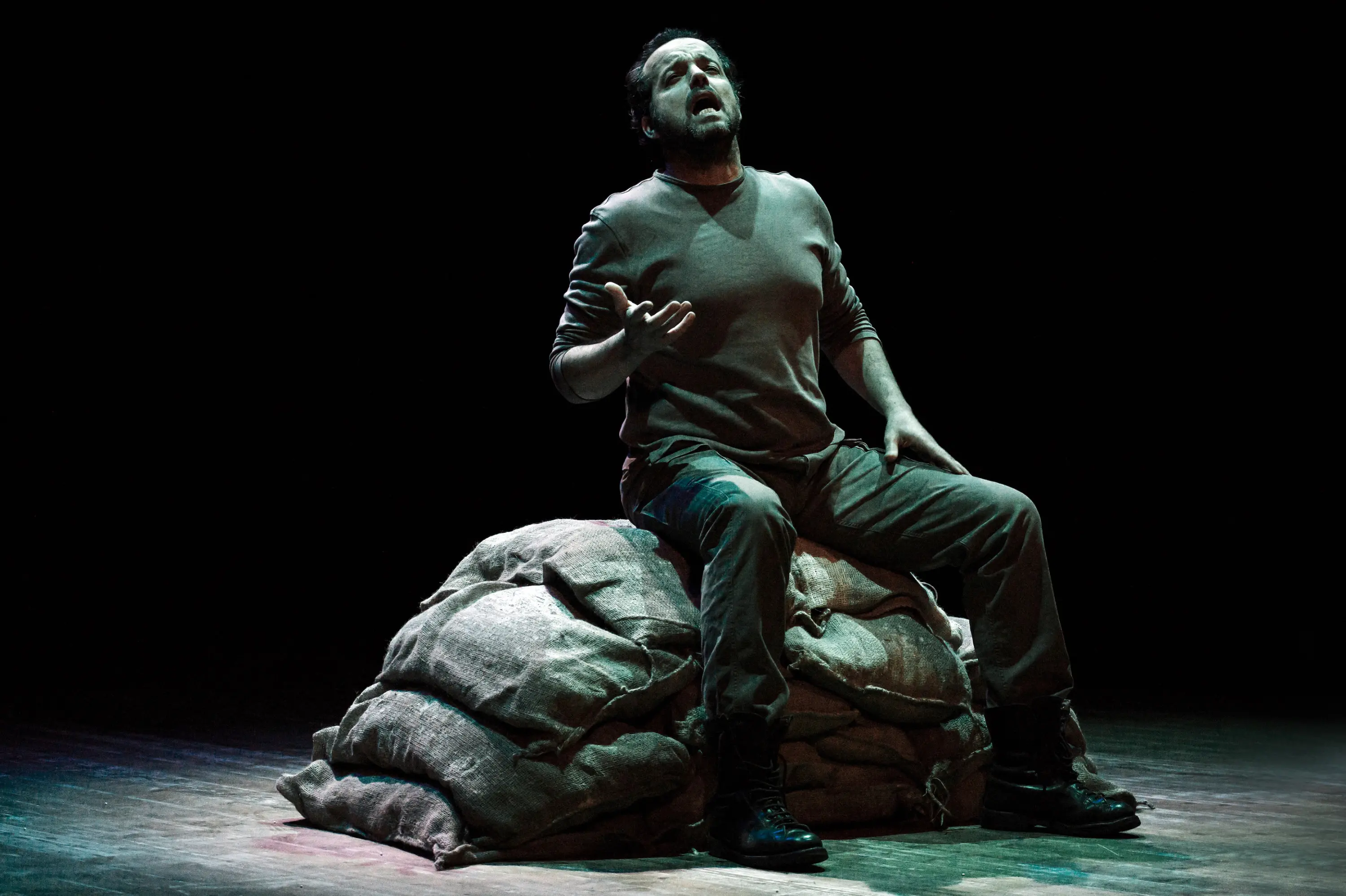 Un attore è seduto su un cumulo di sacchi, con un'espressione intensa. Indossa un maglione grigio e pantaloni scuri, mentre canta o recita con passione in un ambiente buio, illuminato solo da un gioco di luci.