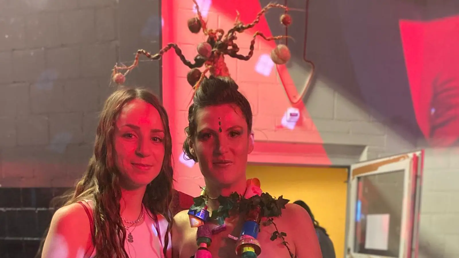 Due donne sorridenti in un ambiente festivo. Una di esse indossa un ornamento fatto di rami e frutta. L'illuminazione è rossa, creando un'atmosfera vibrante.