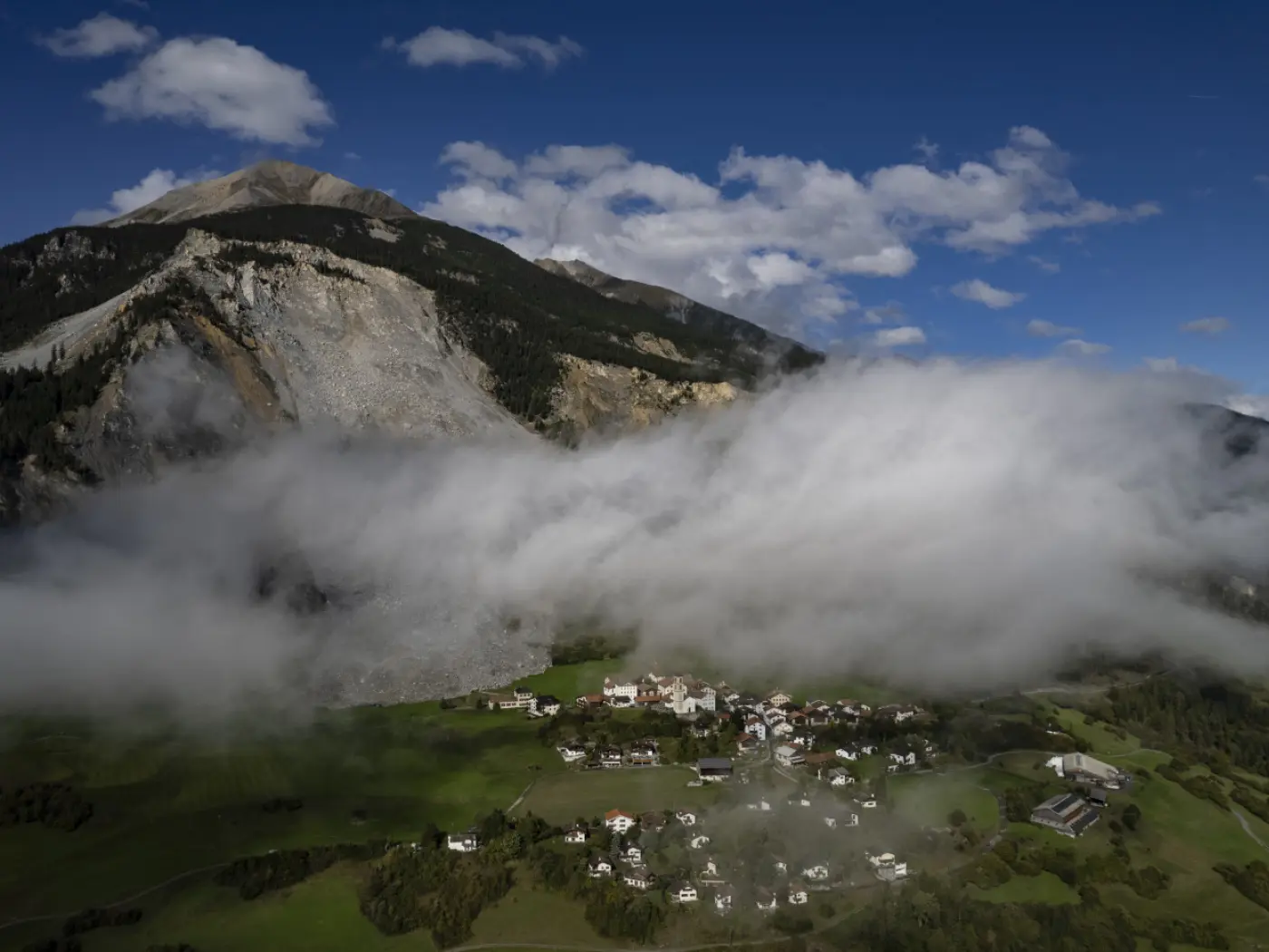 Un villaggio alpino immerso nelle nuvole, con montagne imponenti sullo sfondo. La vegetazione verdeggiante e le case bianche creano un contrasto suggestivo con il cielo blu e le nuvole disperse.