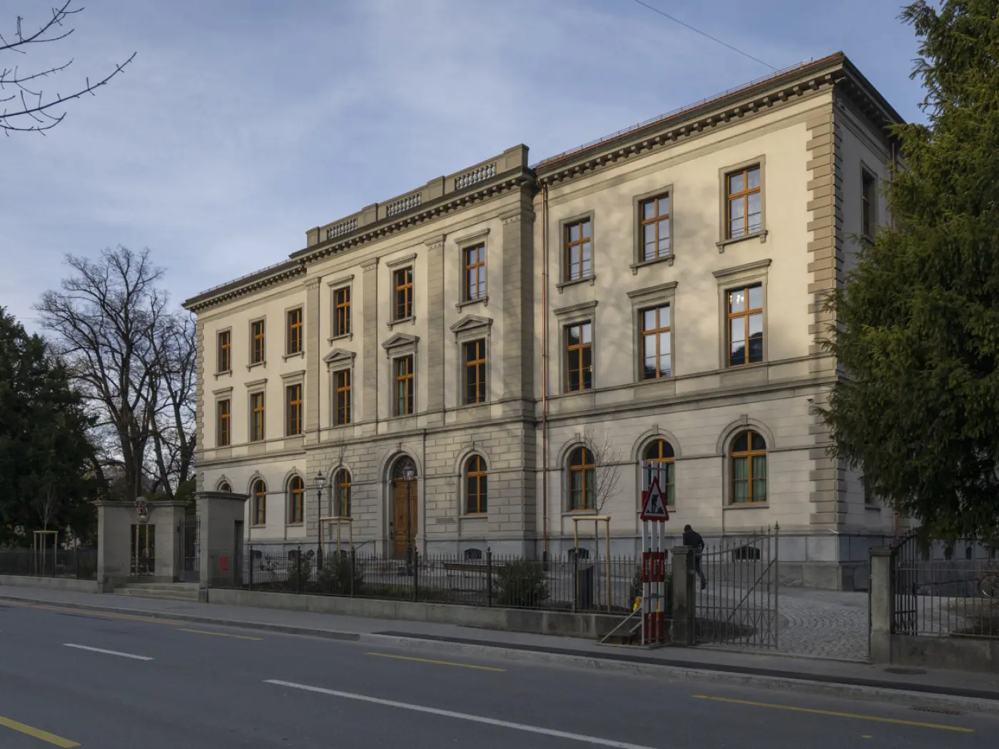 Edificio storico con facciata in stile neoclassico, finestre con persiane beige e dettagli decorativi. La strada si trova di fronte all'edificio, con alberi verdi ai lati.