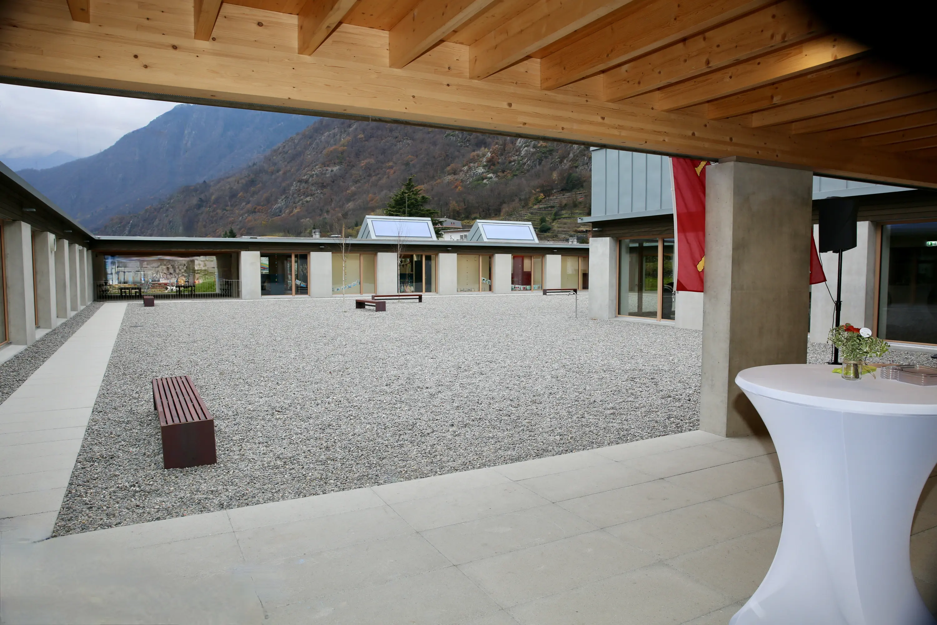 Spazio esterno con pavimentazione in pietra e ghiaia, circondato da edifici moderni con finestre grandi. Si intravedono panchine in legno e una tavola elegante per eventi. Sullo sfondo, si notano le montagne.