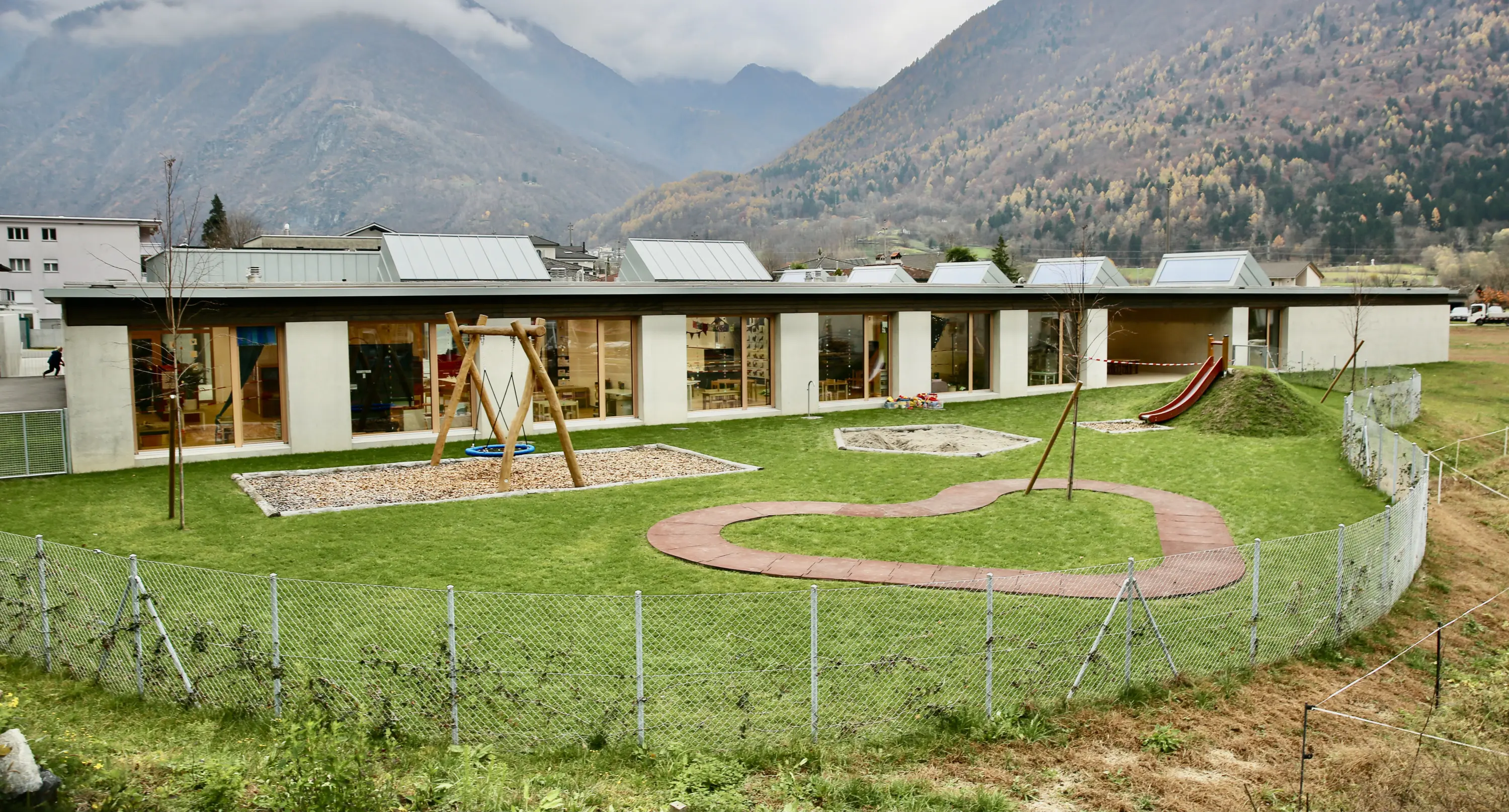 Un edificio moderno e luminoso con ampie finestre, circondato da un'area verde curata. Un giardino per bambini presenta un'altalena e uno scivolo su un manto erboso, mentre il paesaggio montano fa da sfondo.