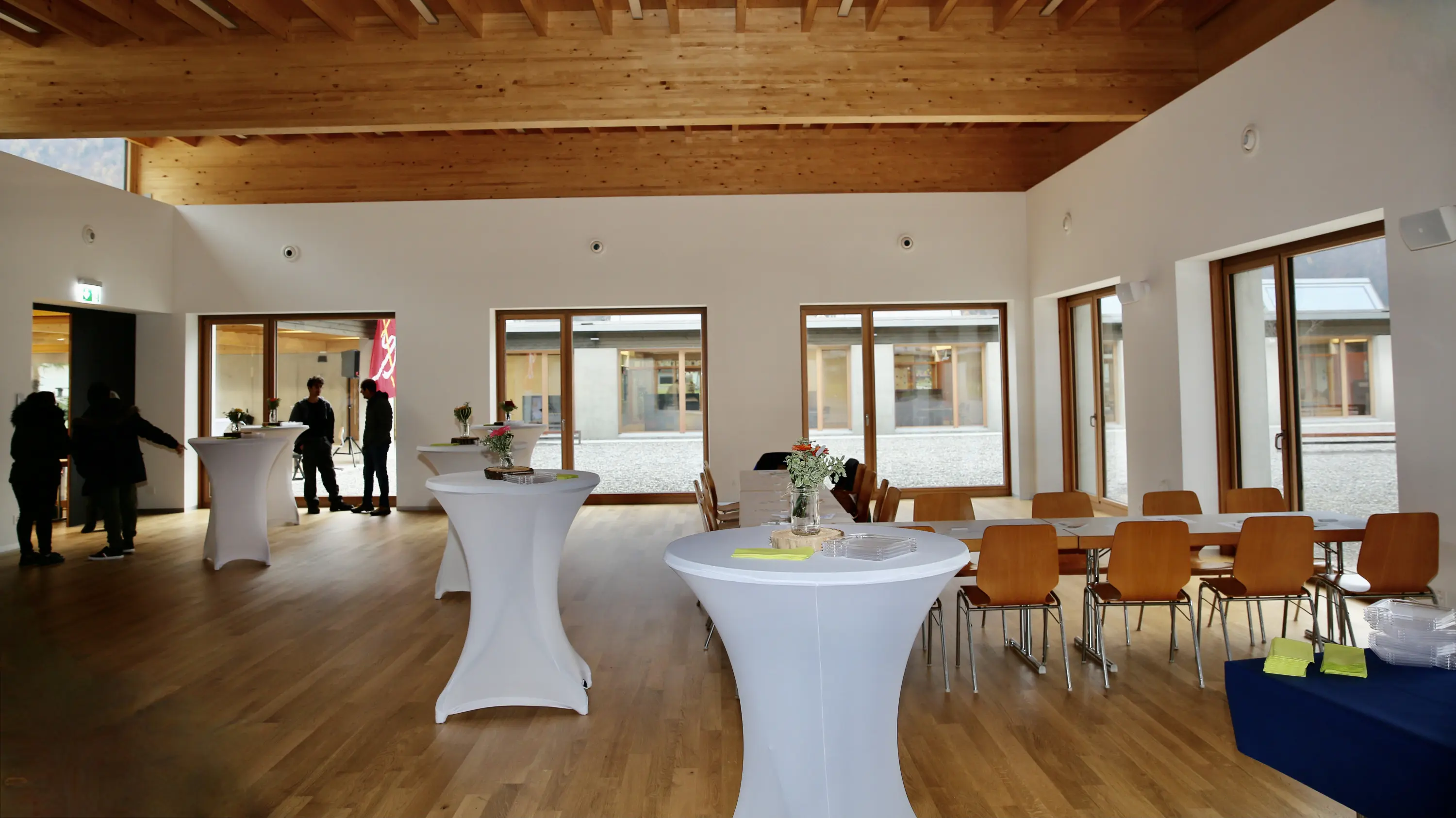 Ambiente interno di un moderno spazio per eventi, con pavimenti in legno e grandi finestre. Sono presenti tavoli alti bianchi e sedie di legno. Alcune persone interagiscono sullo sfondo.