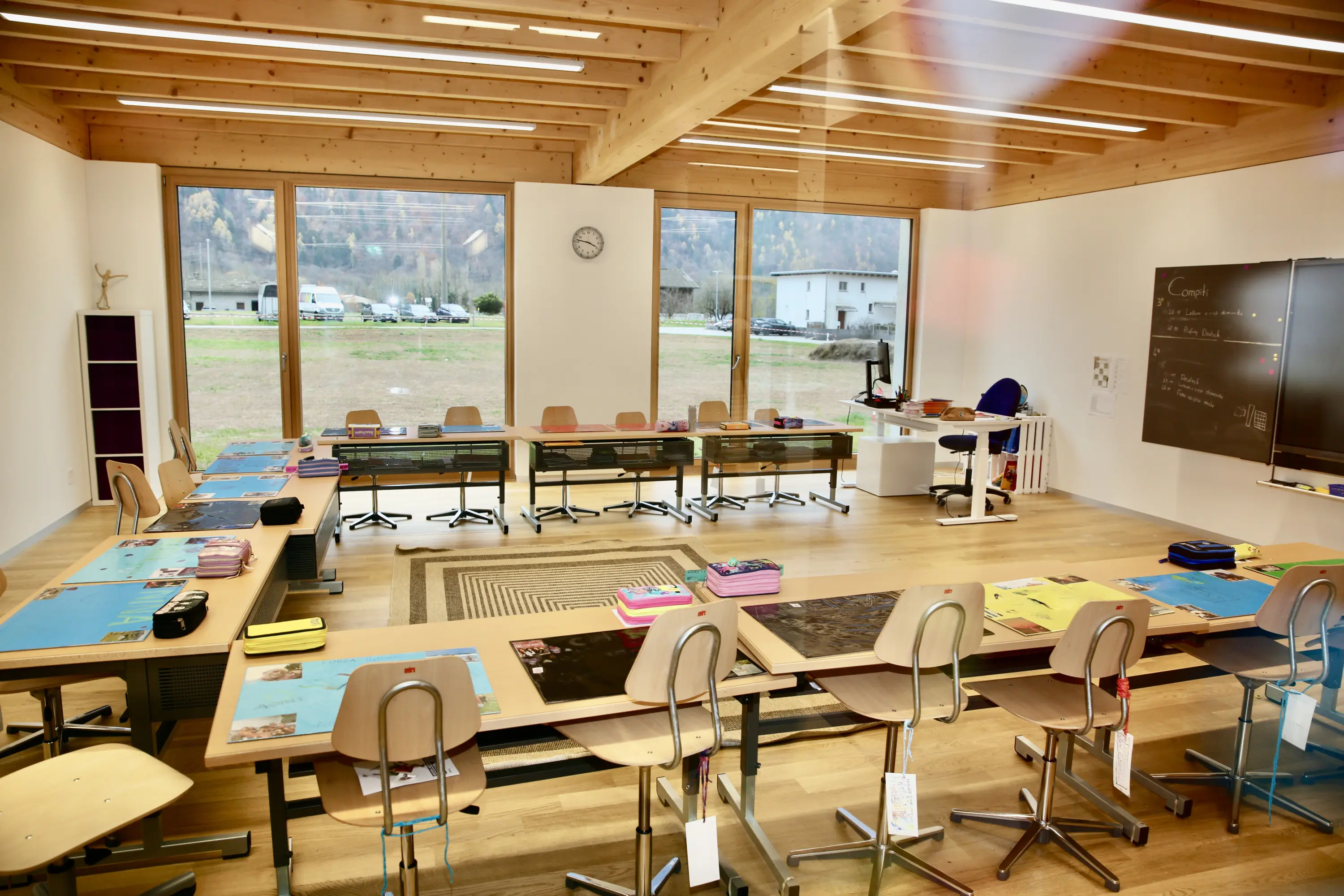 Aula moderna con pareti bianche e grandi finestre che mostrano un paesaggio esterno. I banchi sono disposti ordinatamente con materiali scolastici sopra. Una lavagna nera è visibile su un lato. Un orologio è appeso a una pare
