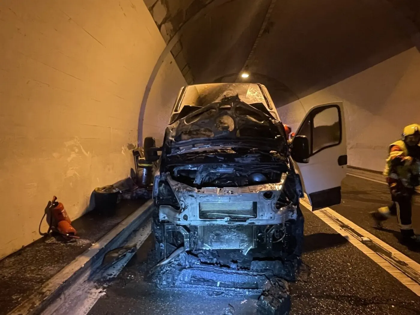 Furgone danneggiato all'interno di un tunnel, con il motore in fiamme e i vigili del fuoco in azione. Una bombola di estintore è visibile a sinistra. L'illuminazione è fioca, con riflessi