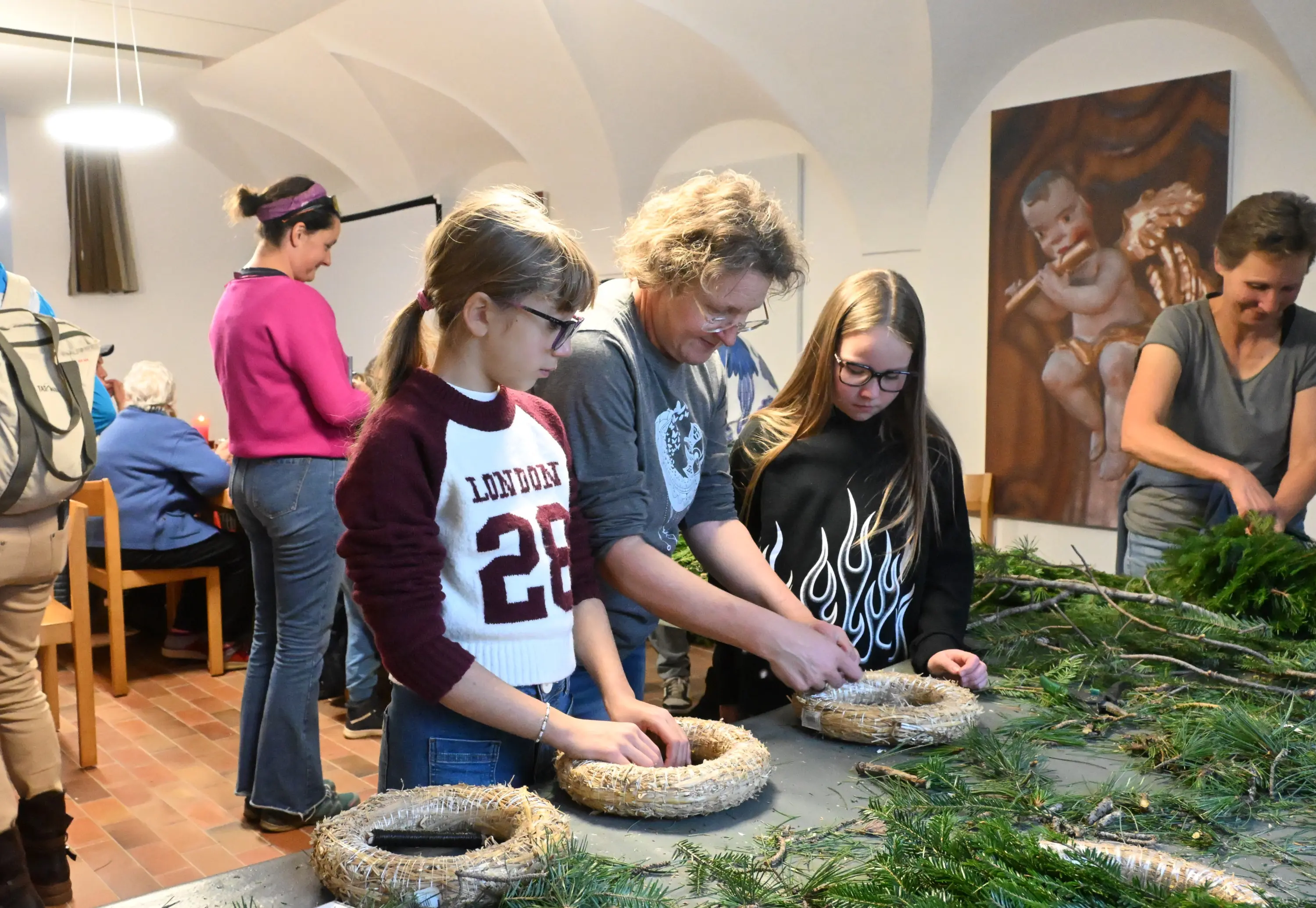Un gruppo di persone in un ambiente interno sta creando ghirlande natalizie. Due ragazze e un adulto lavorano insieme su un tavolo, mentre altre persone sono visibili sullo sfondo. Elementi naturali come rami e decorazioni sono sparsi sul tavolo.