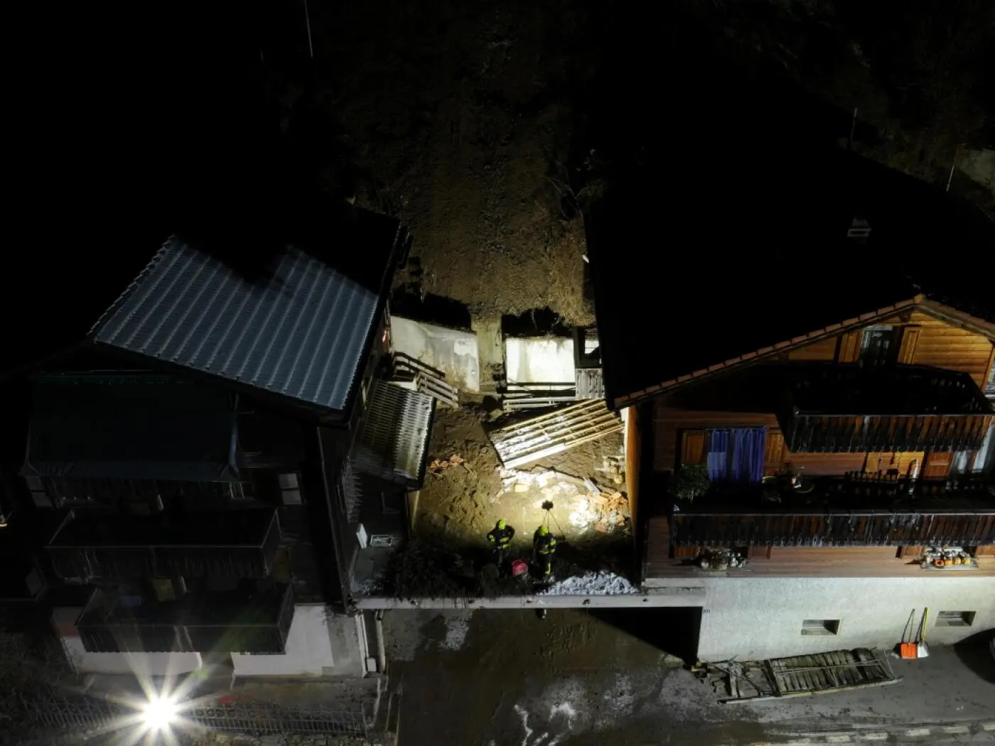 Due edifici in legno illuminati di notte, con un’area di costruzione o di attività in corso tra di essi. Due persone sono visibili al lavoro in mezzo a materiale da costruzione e detriti. La scena è avvolta nell'oscurità, con luc