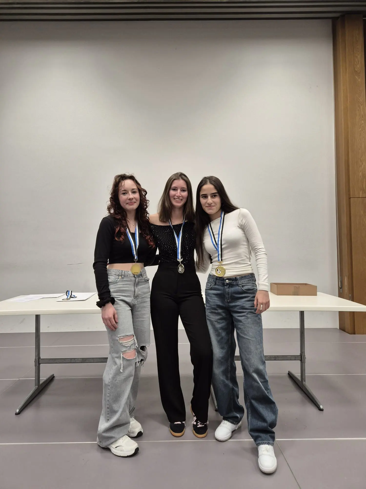 Tre ragazze sorridenti indossano medaglie d'oro e posano insieme davanti a un tavolo. Due hanno i capelli ricci e mossi, mentre la terza ha i capelli lisci. Indossano abiti diversi, con stili casual.