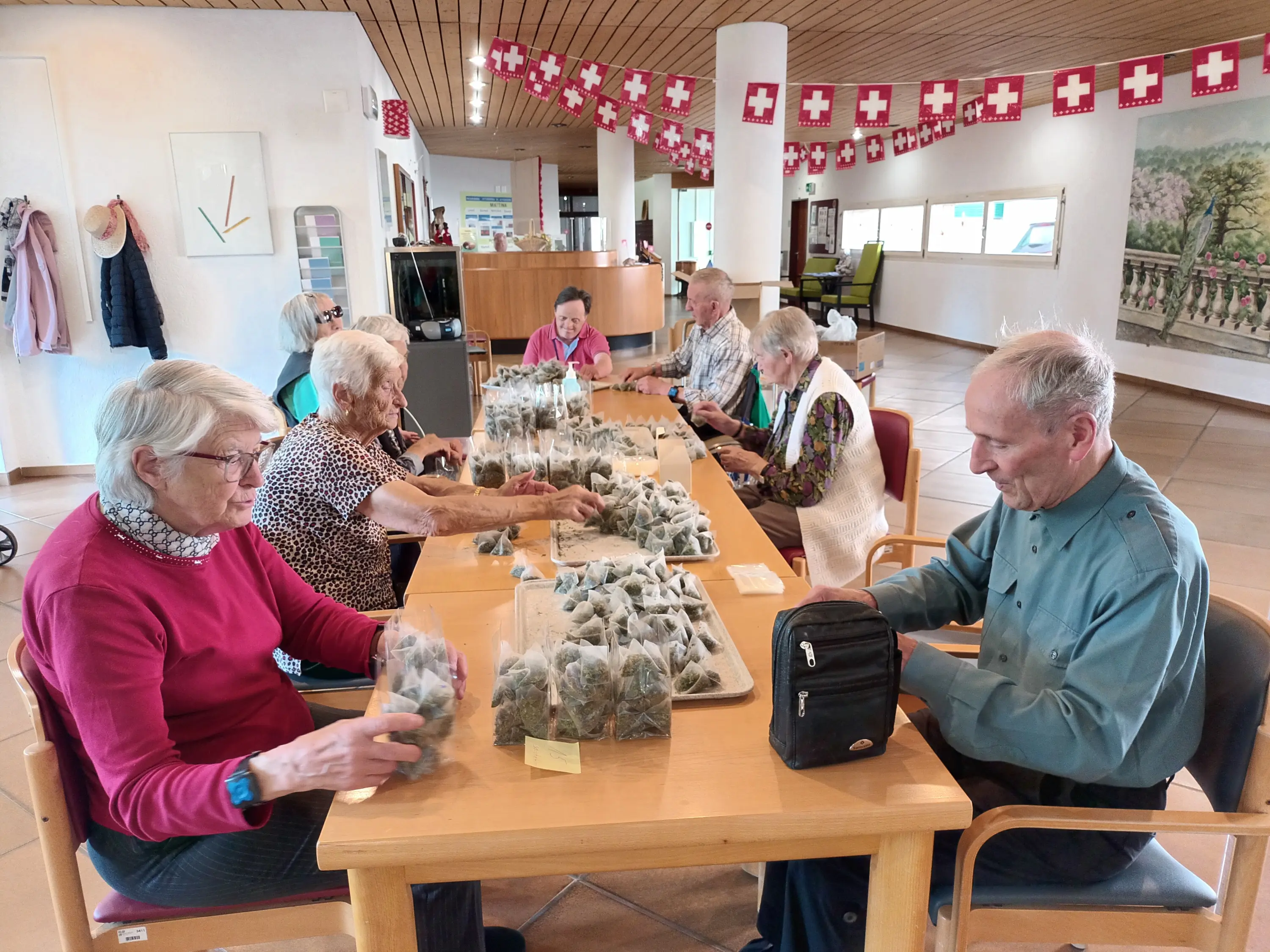 Un gruppo di anziani in un centro di assistenza, seduti attorno a un tavolo, mentre confezionano sacchetti. Alle pareti sono appesi festoni svizzeri. L'atmosfera è di attività e socializzazione.