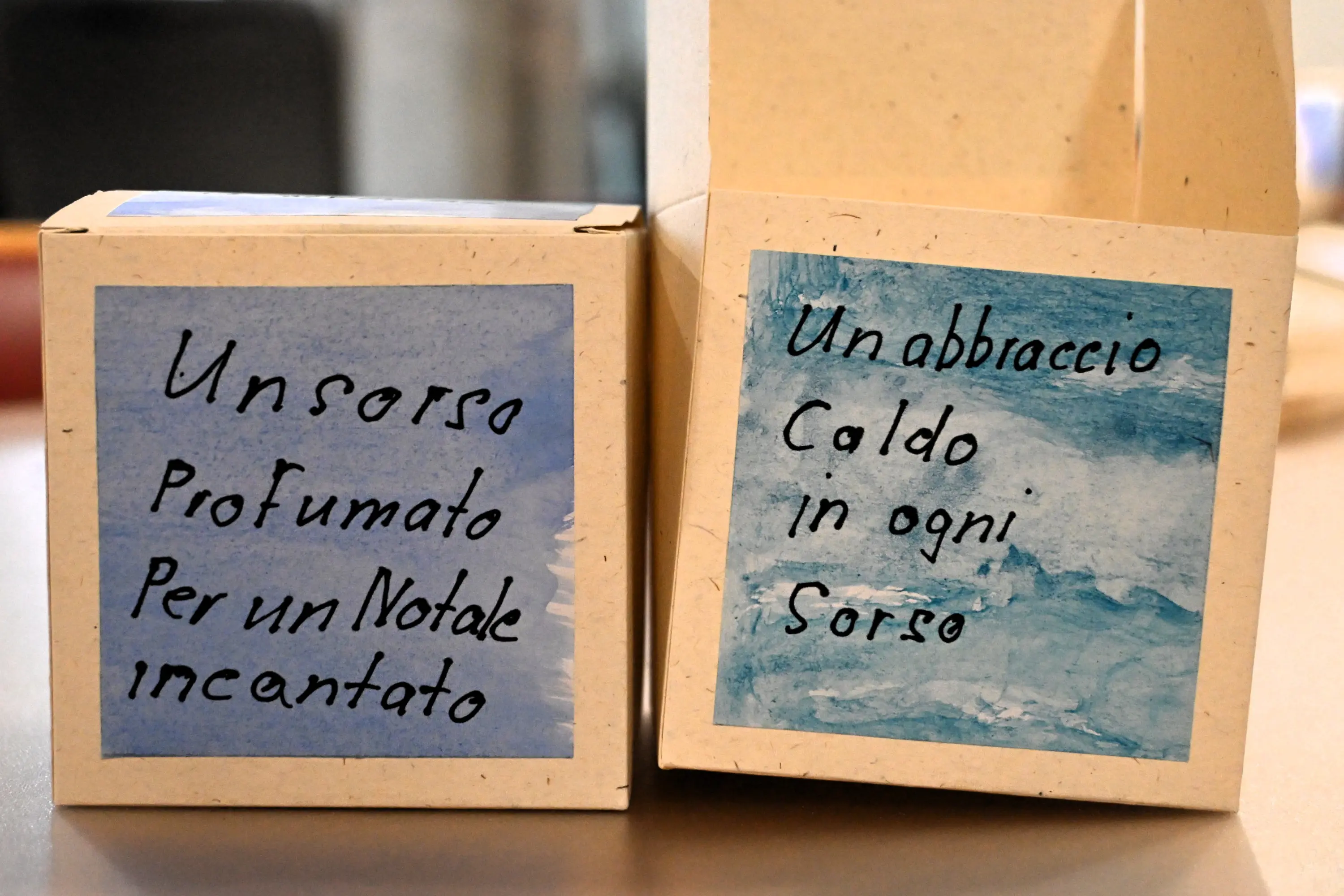 Due scatole di cartone con scritte artistiche. Una presenta la frase "Un sorso profumato per un Natale incantato" e l'altra "Un abbraccio caldo in ogni sorso".