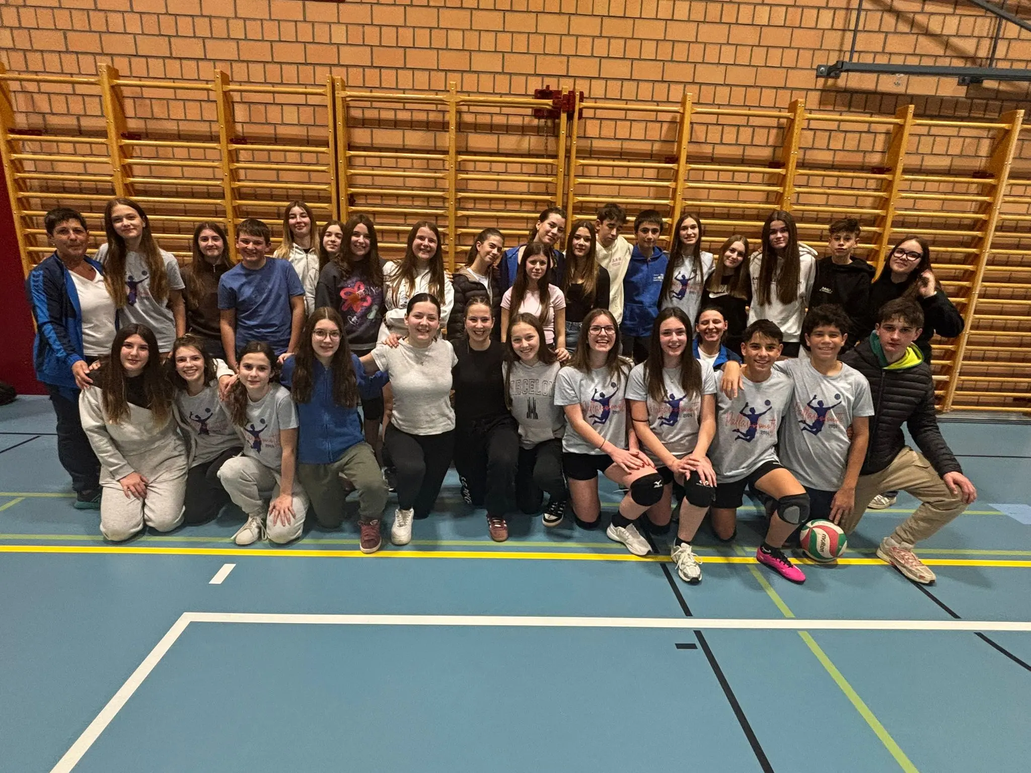 Gruppo di giovani atleti in una palestra, sorridenti e in tuta sportiva, posano insieme per una foto di squadra. Alcuni indossano magliette con un motivo di pallavolo.