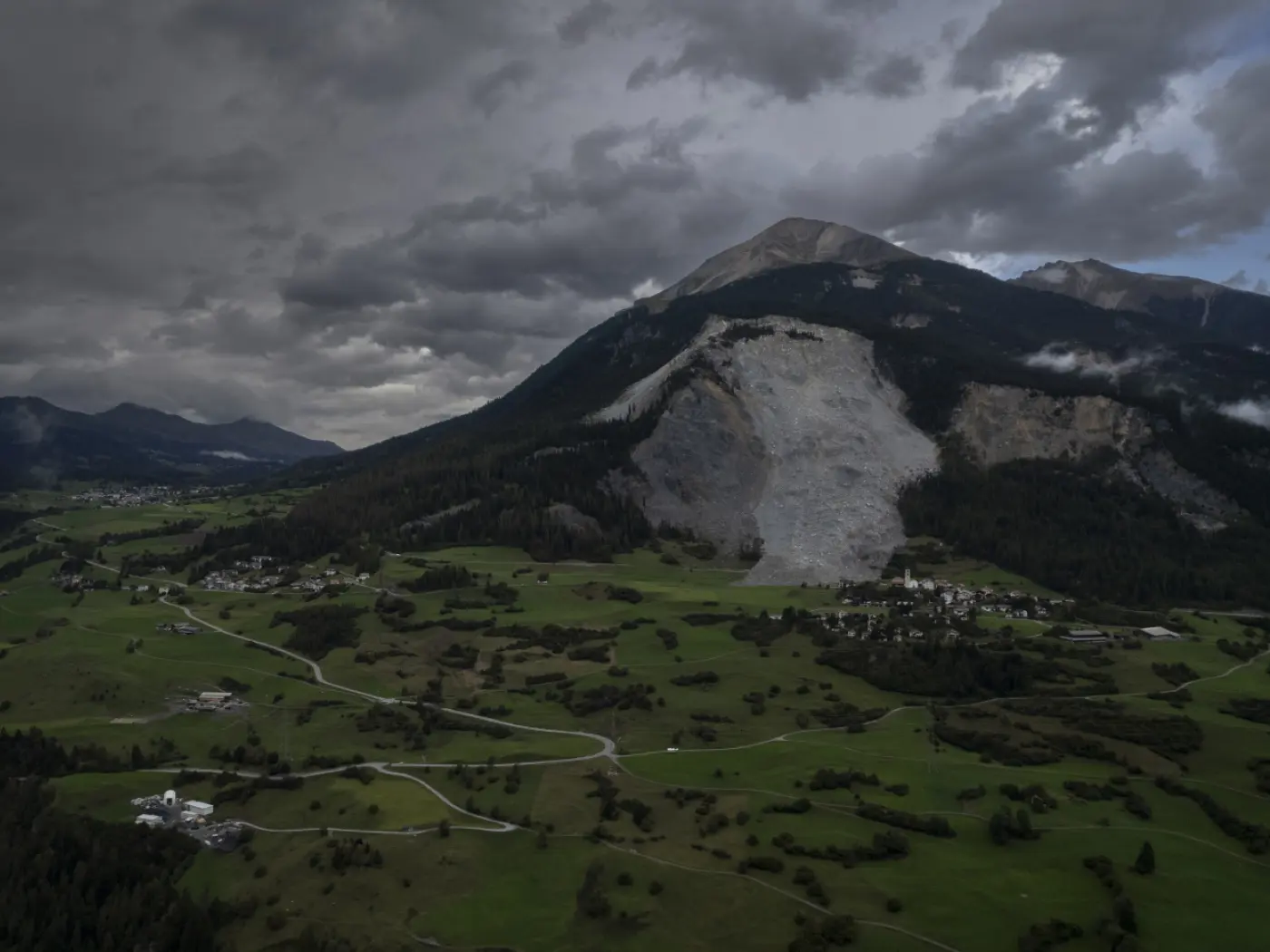 Un paesaggio montano con una grande frana visibile sulla parete di una montagna. Nuvole scure coprono il cielo, mentre la valle sottostante presenta prati verdi e piccoli insediamenti.