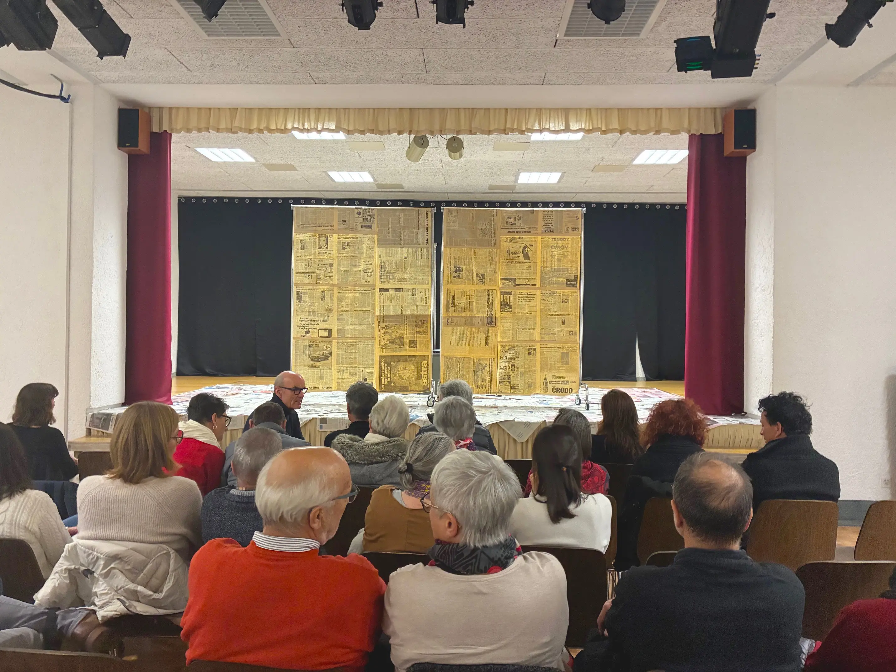 Un gruppo di persone sedute su panche, che osserva un palcoscenico coperto da una struttura di cartone con articoli di giornale. Il palco è allestito per uno spettacolo e le tende sono chiuse.