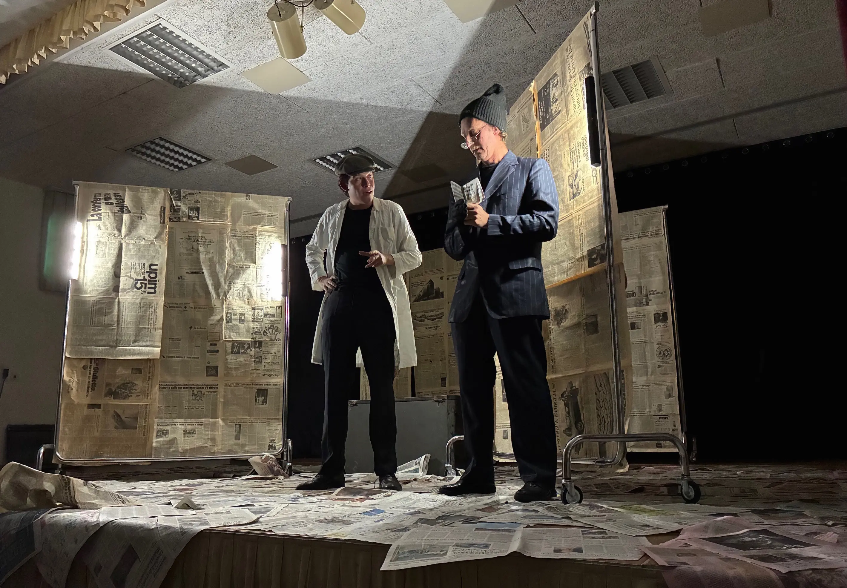 Due persone sono in scena, circondate da pannelli di cartone e giornali sparsi a terra. Una persona indossa un camice bianco e occhiali, mentre l'altra è in giacca e berretto. Entrambi sembrano discutere.