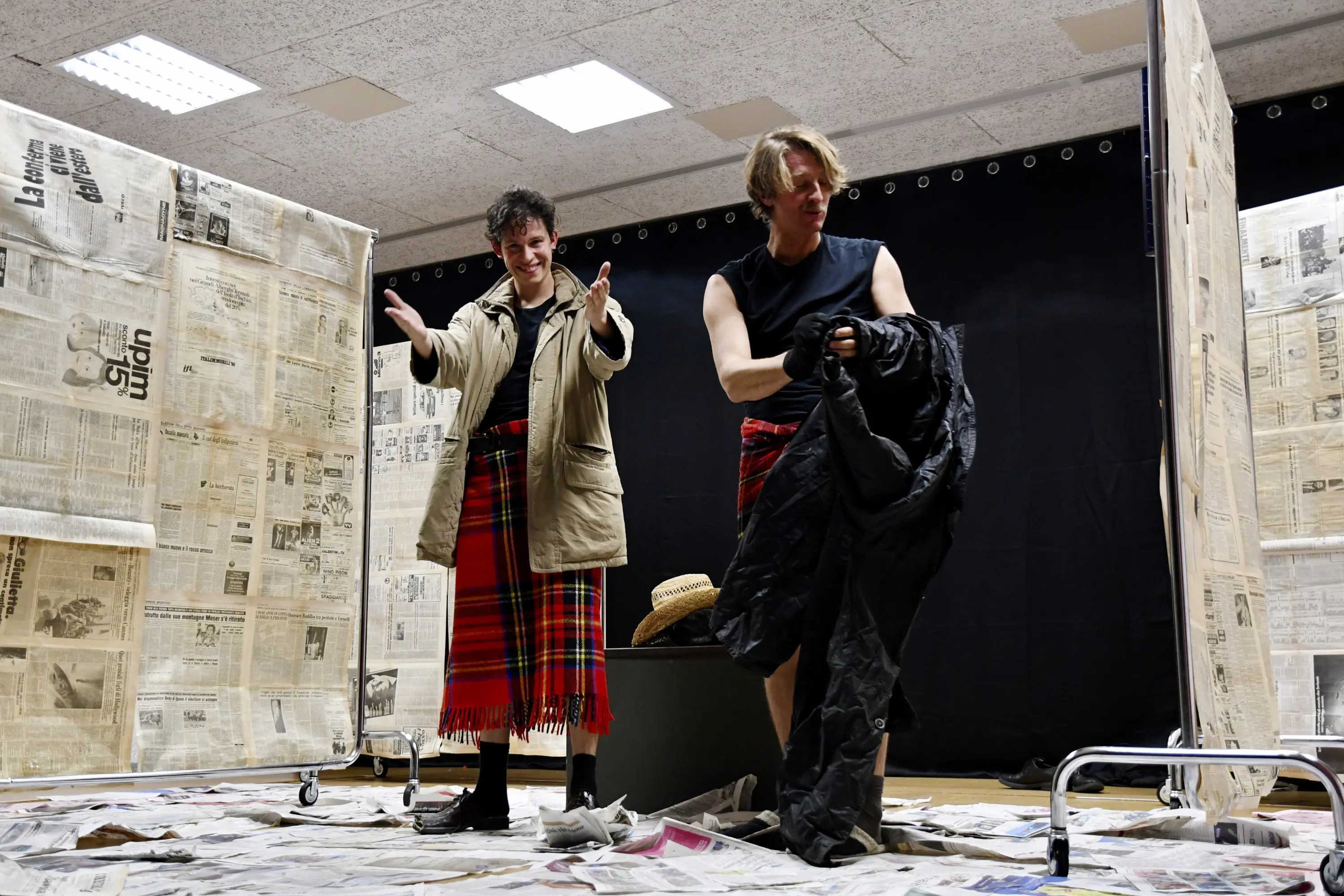 Due attori sul palco, uno indossa una giacca beige e una gonna scozzese, mentre l'altro è in maglietta nera e pantaloni. Sono circondati da fogli di giornale e pareti di cartone. L'atmosfera sembra essere di un