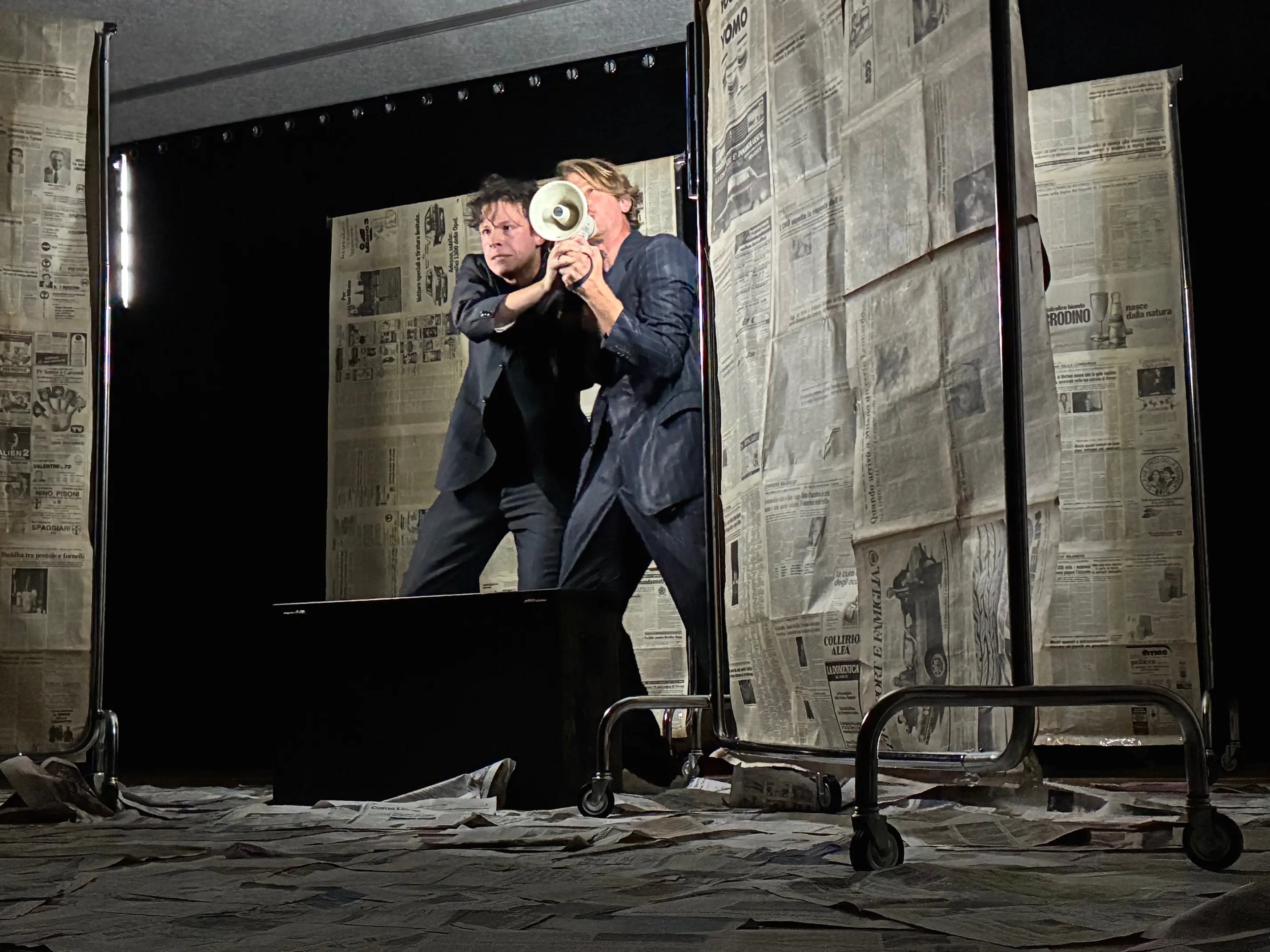 Due attori in abiti scuri si abbracciano mentre puntano un megafono verso il pubblico. Sono circondati da pannelli di cartone ricoperti di giornali e un pavimento disseminato di carta.
