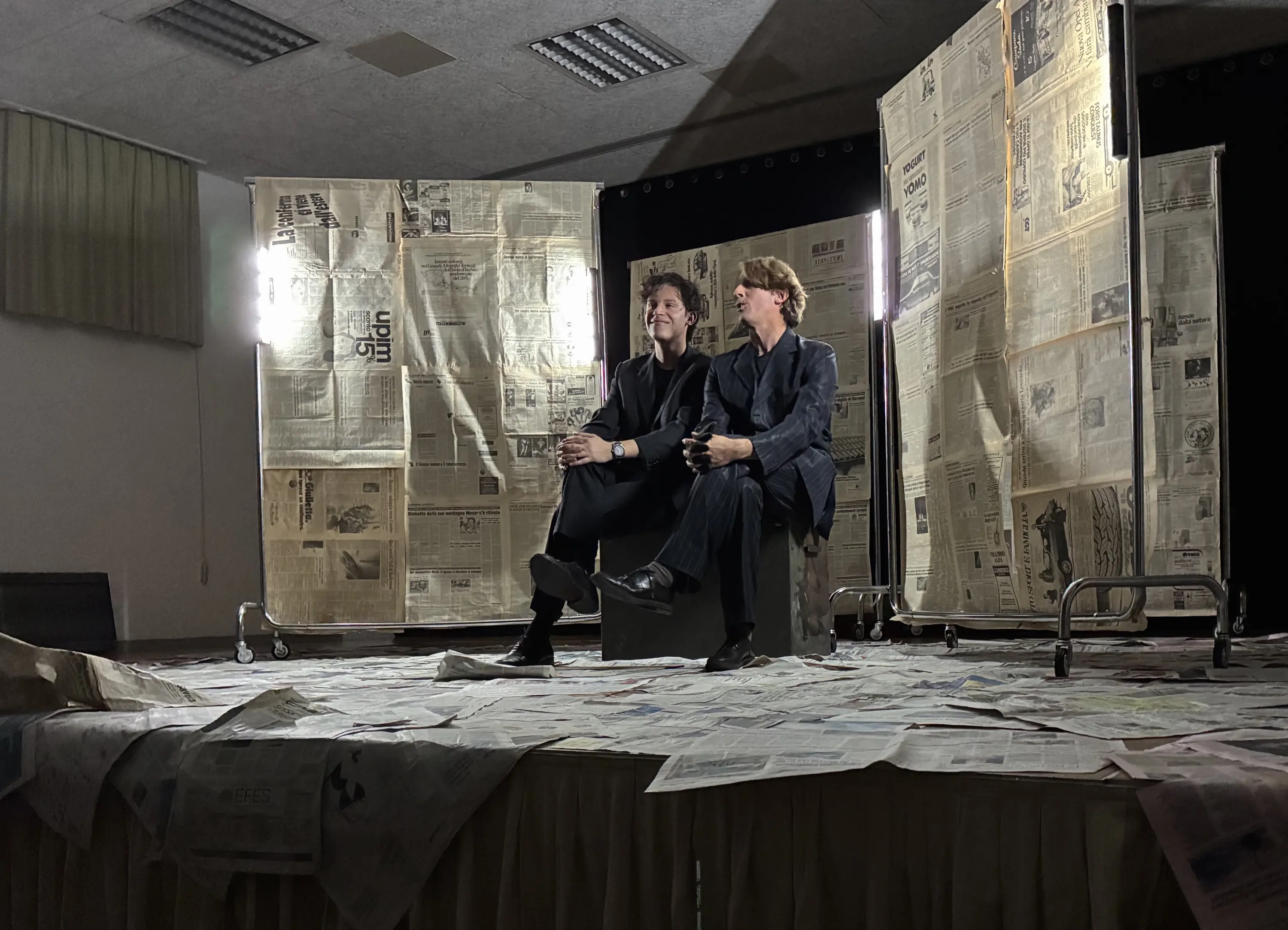 Due uomini seduti su una sedia in un ambiente con pareti di carta da giornale. I volti dei due uomini sorridono, mentre sono circondati da una scenografia semplice ma creativa.