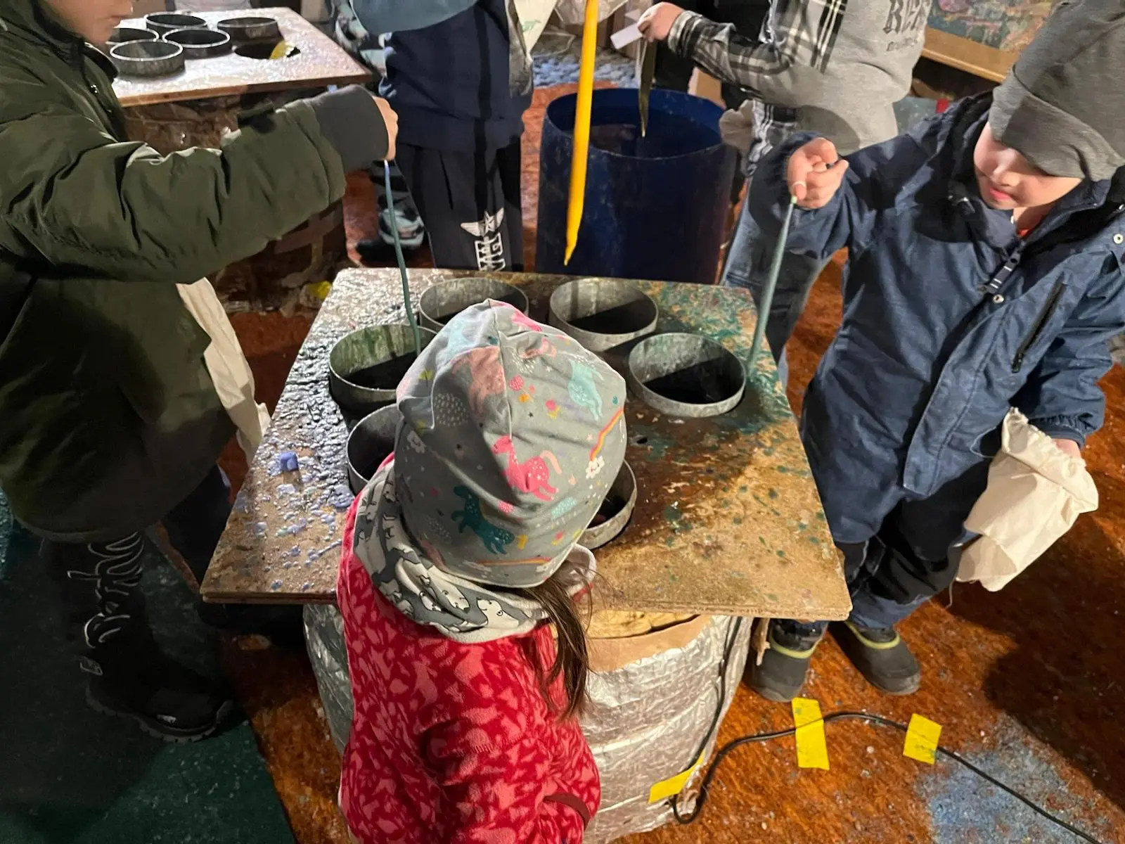 Bambini che utilizzano recipienti per attività manuali in un ambiente interno. Alcuni hanno indossato cappelli e sciarpe, mentre si concentrano su ciò che stanno facendo.