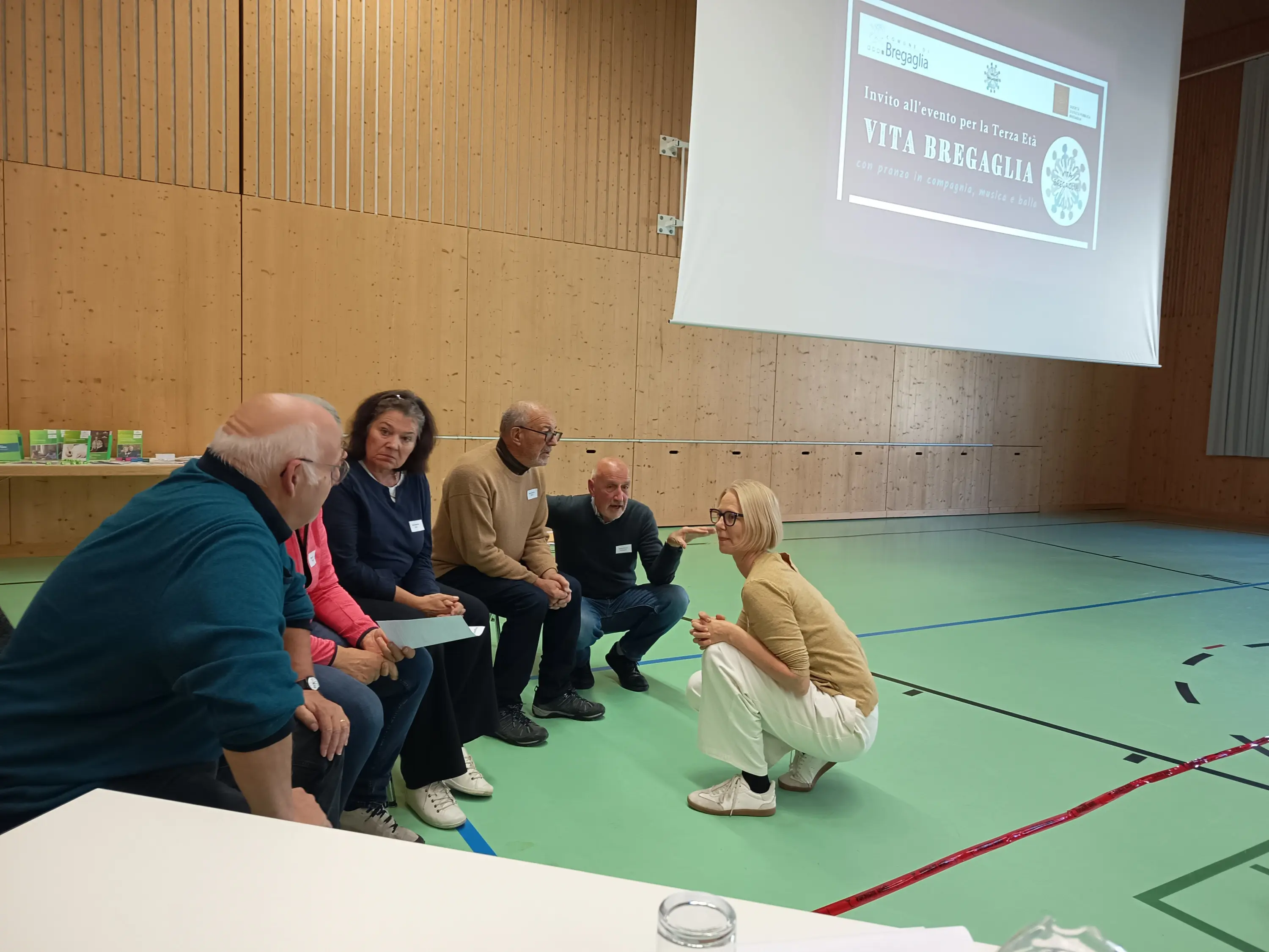 Un gruppo di sei persone sedute su una panchina discusso davanti a una donna in piedi che li ascolta attentamente. Sullo sfondo si vede un grande schermo con del testo. L'ambiente è una palestra con pareti di legno.