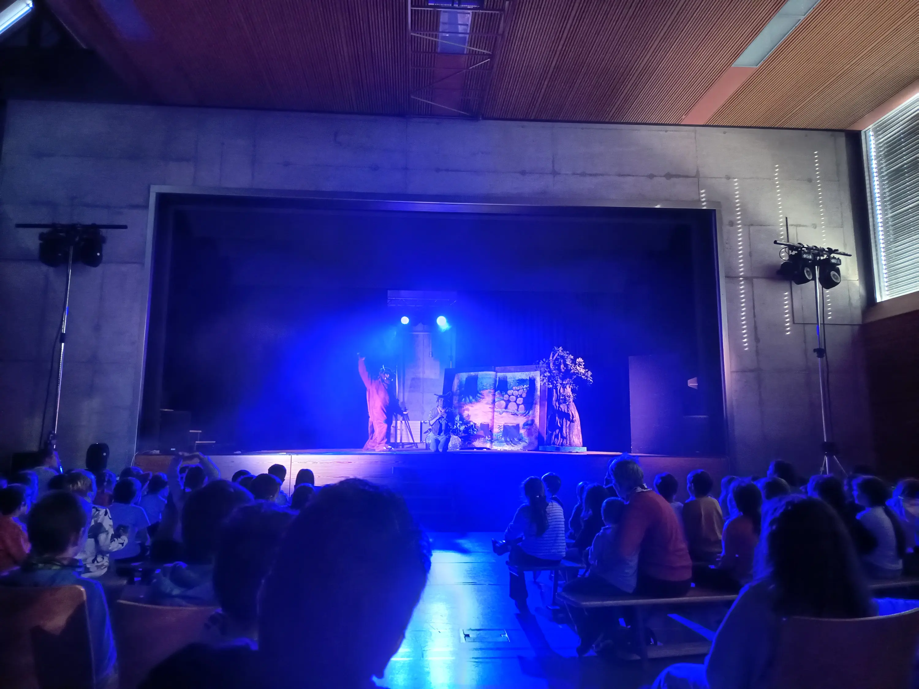 Un palcoscenico illuminato da luci blu, con due performer in costumi artistici che danzano. Un pubblico vario osserva con attenzione. Sullo sfondo, un'ambientazione scenografica è visibile.