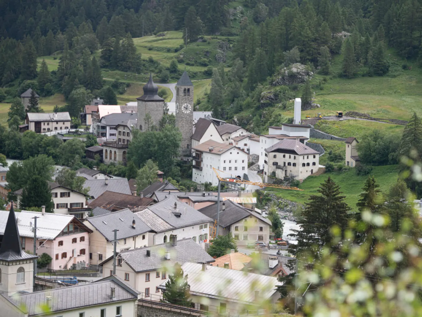 Panorama di un piccolo villaggio montano con case tradizionali e una torre campanaria. Sullo sfondo si possono vedere alberi verdi e colline.