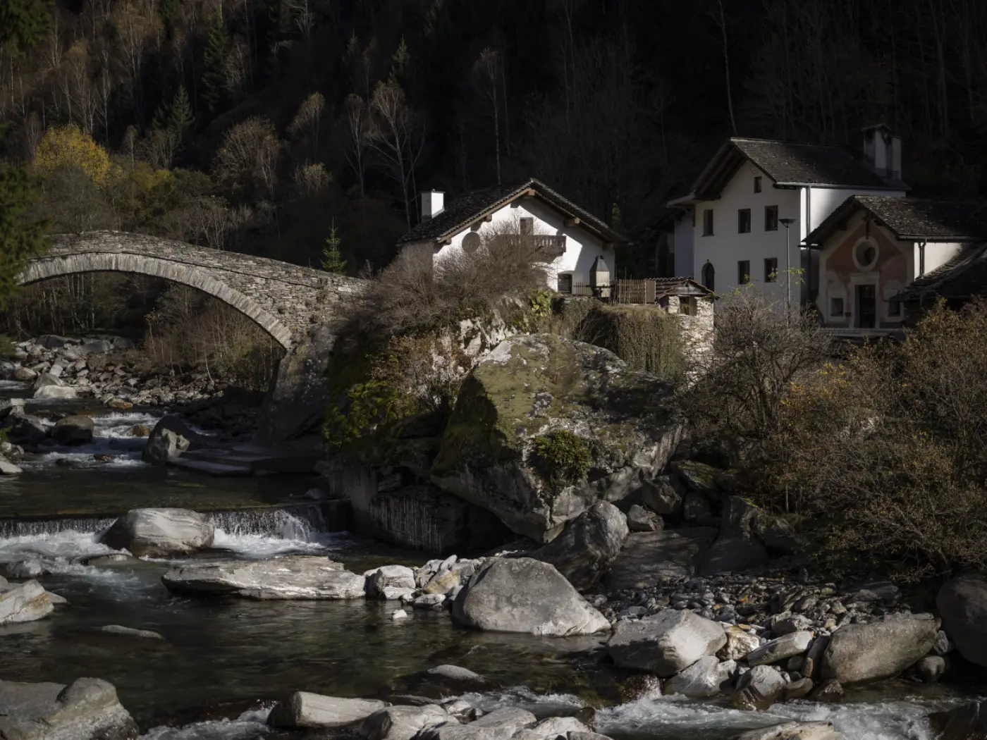Un ponte di pietra attraversa un fiume, con case tradizionali in lontananza circondate da alberi autunnali. Rocce di diverse dimensioni si trovano nel fiume.