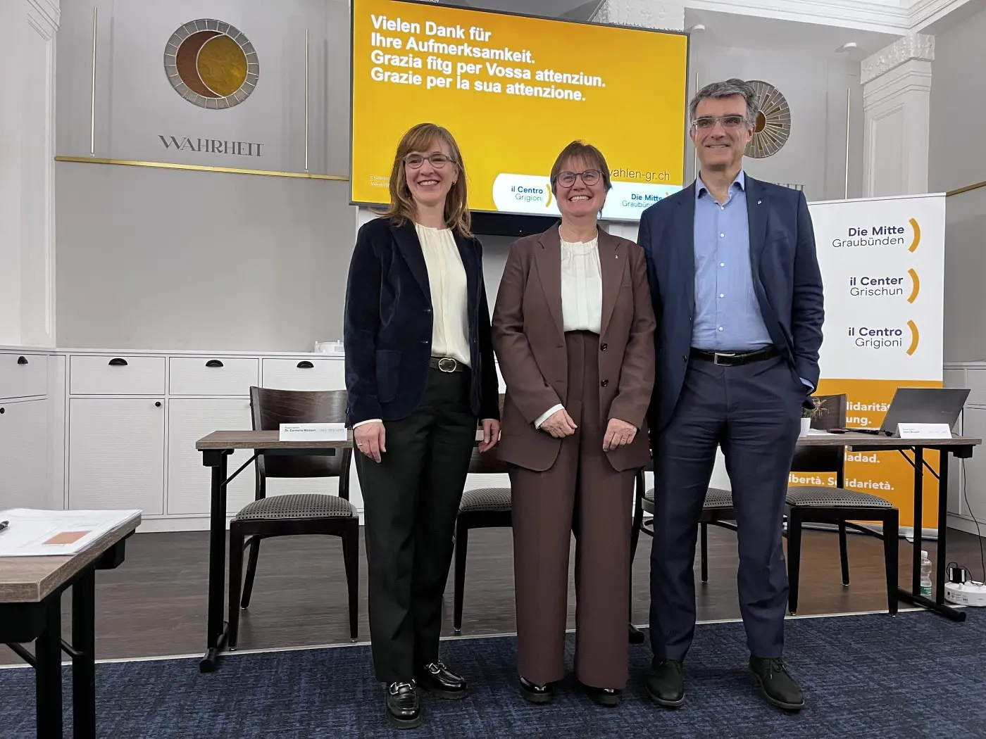 Tre persone sorridono all'interno di una sala conferenze. Sullo sfondo, un grande schermo mostra un messaggio di ringraziamento in tedesco e italiano. I partecipanti indossano abiti formali.