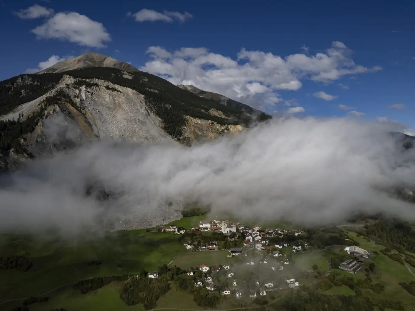Una vista aerea di un villaggio montano circondato da nuvole basse, con una grande montagna sullo sfondo e un cielo parzialmente nuvoloso.