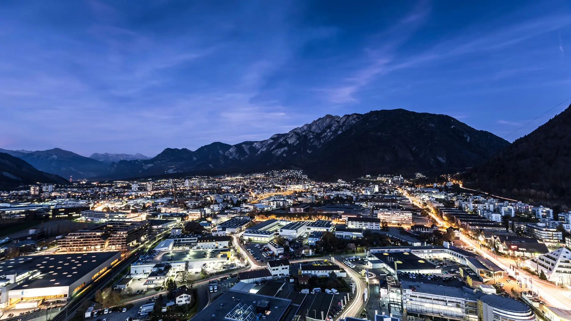 Veduta notturna di una città illuminata, circondata da montagne. Le luci delle strade e degli edifici si riflettono nel cielo blu scuro, creando un'at