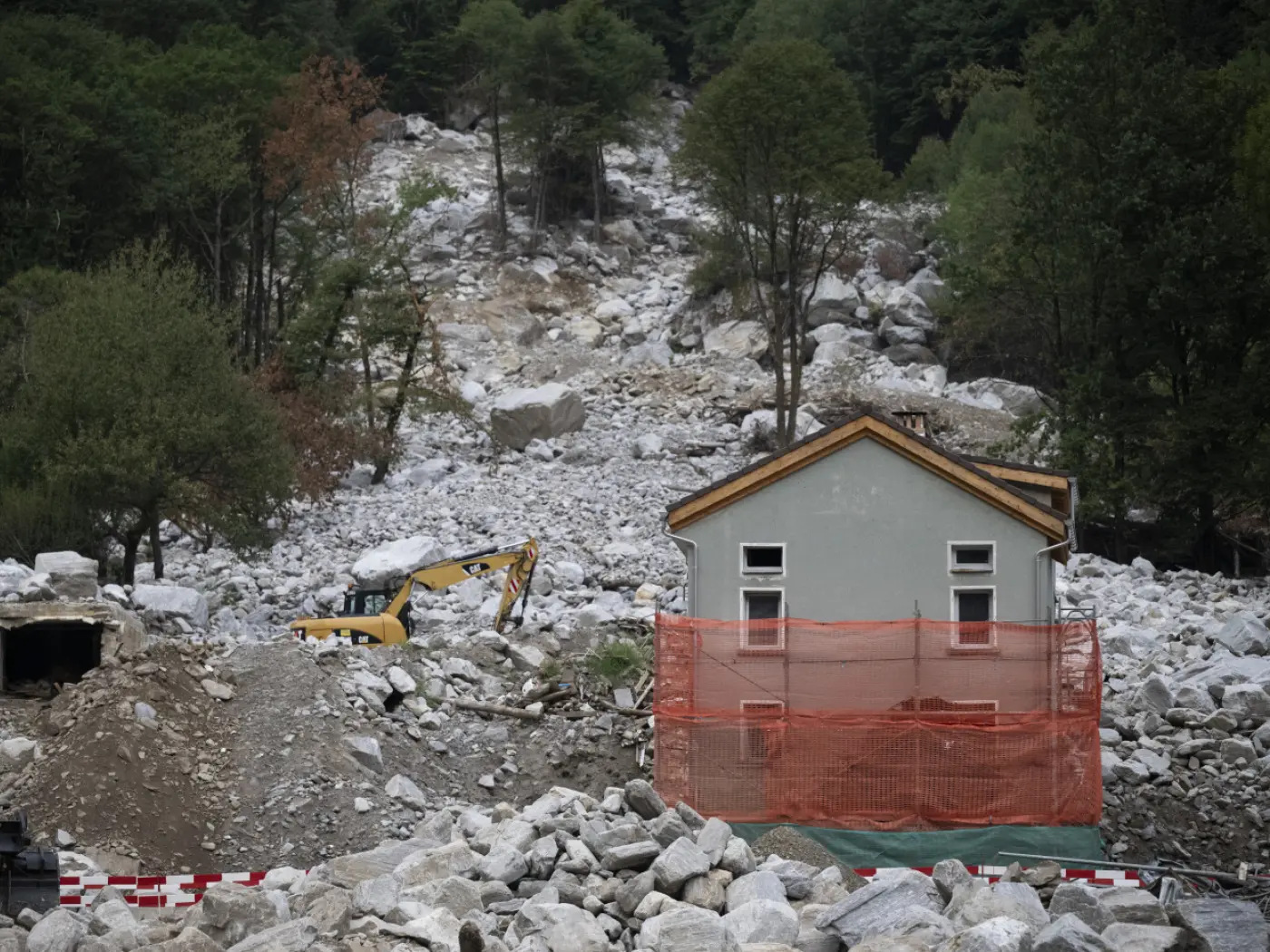 Una casa parzialmente sepolta da una frana di rocce e detriti. Sullo sfondo si vede un escavatore giallo e la vegetazione circostante. Una