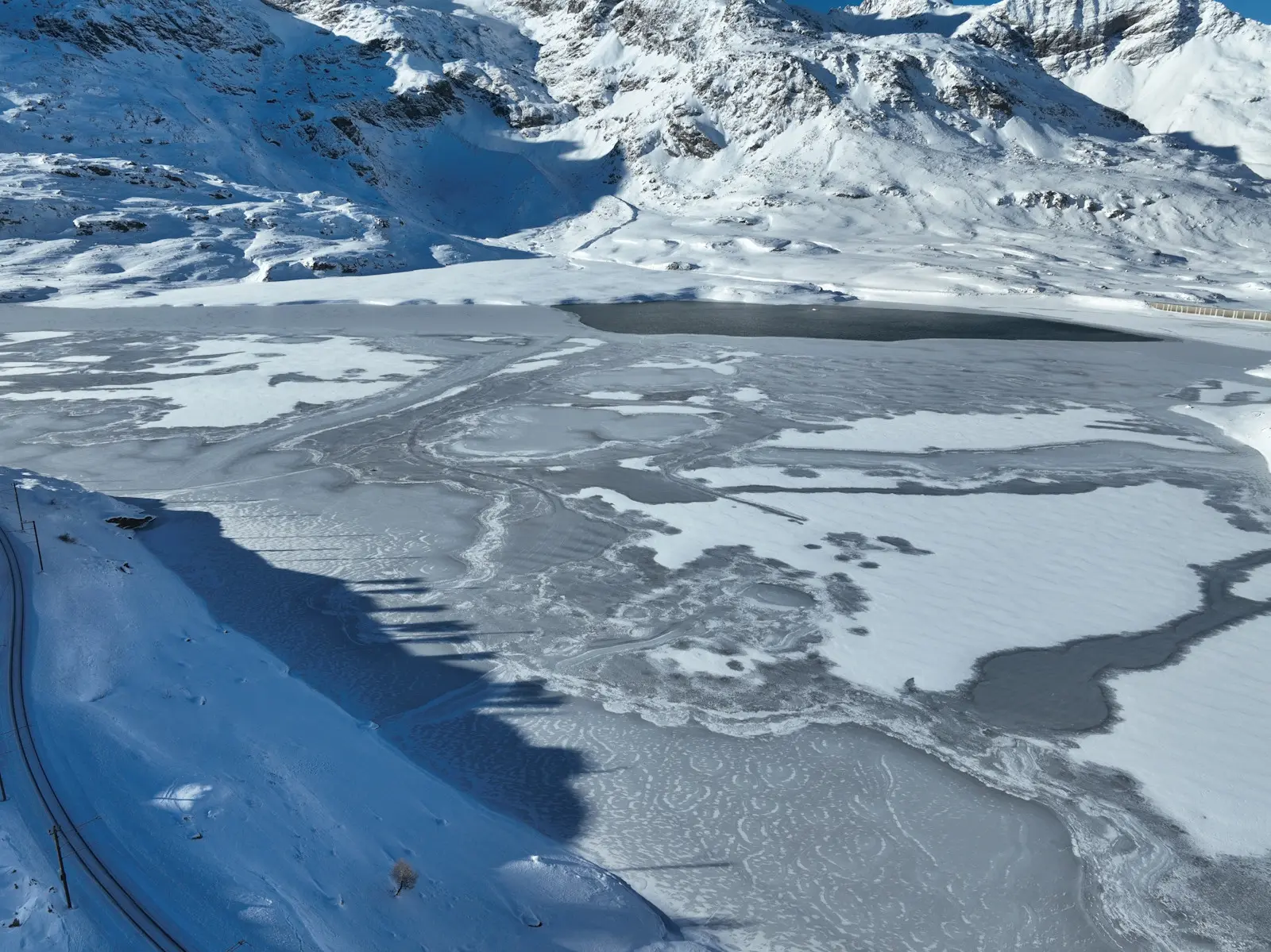 Paesaggio montano innevato con un lago parzialmente ghiacciato. Le montagne circostanti sono coperte di neve, creando un contrasto suggestivo con il ghiaccio