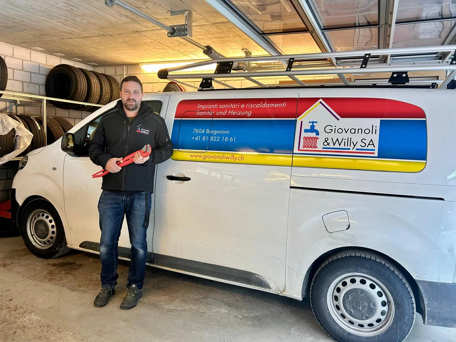 Uomo sorridente con giacca nera tiene un attrezzo rosso accanto a un furgone bianco con il logo di "Giovanoli & Willy SA". Sullo sf