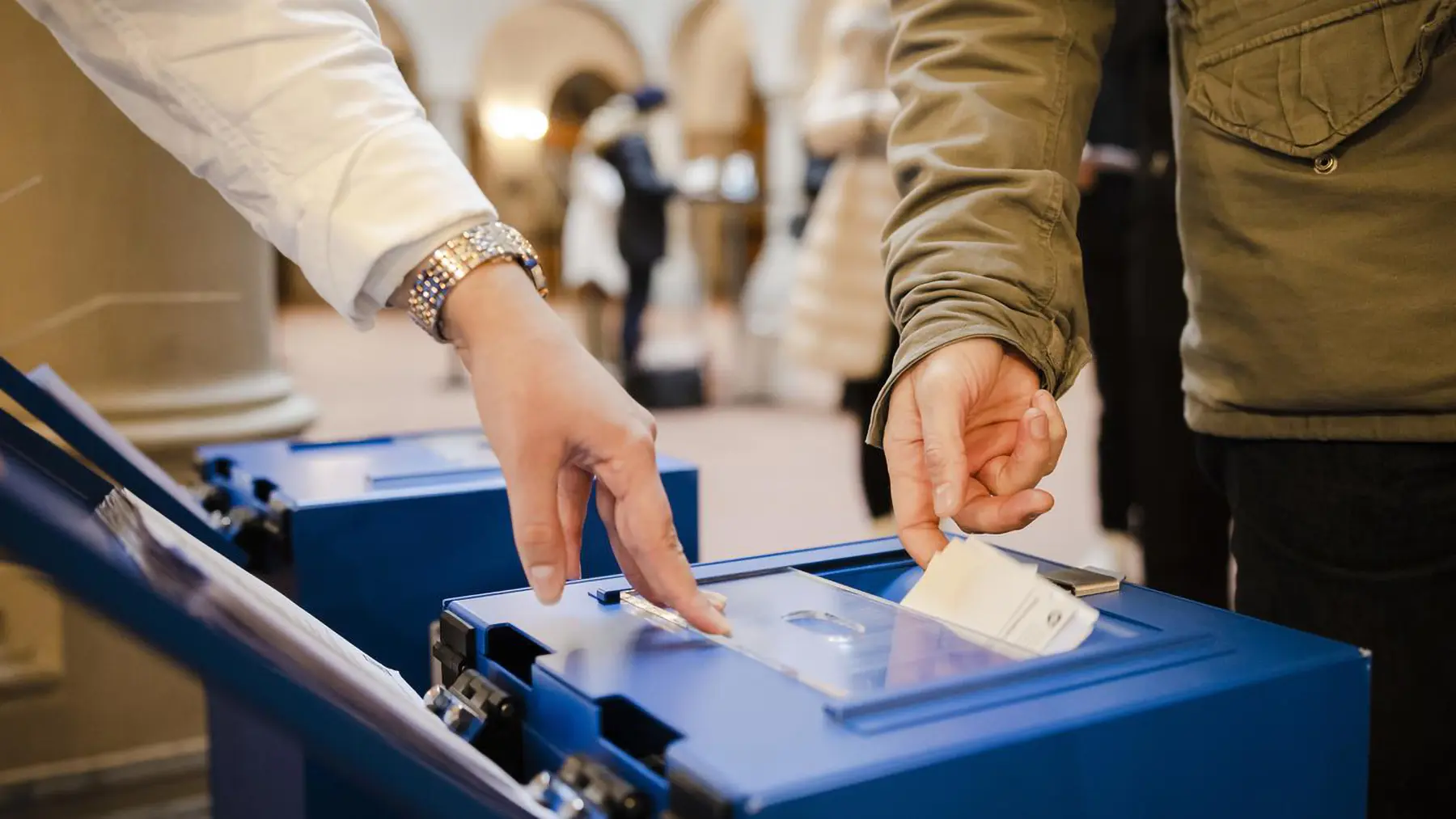 Due mani inseriscono una scheda elettorale in una urna blu. Sullo sfondo si vedono altre persone che partecipano al voto.