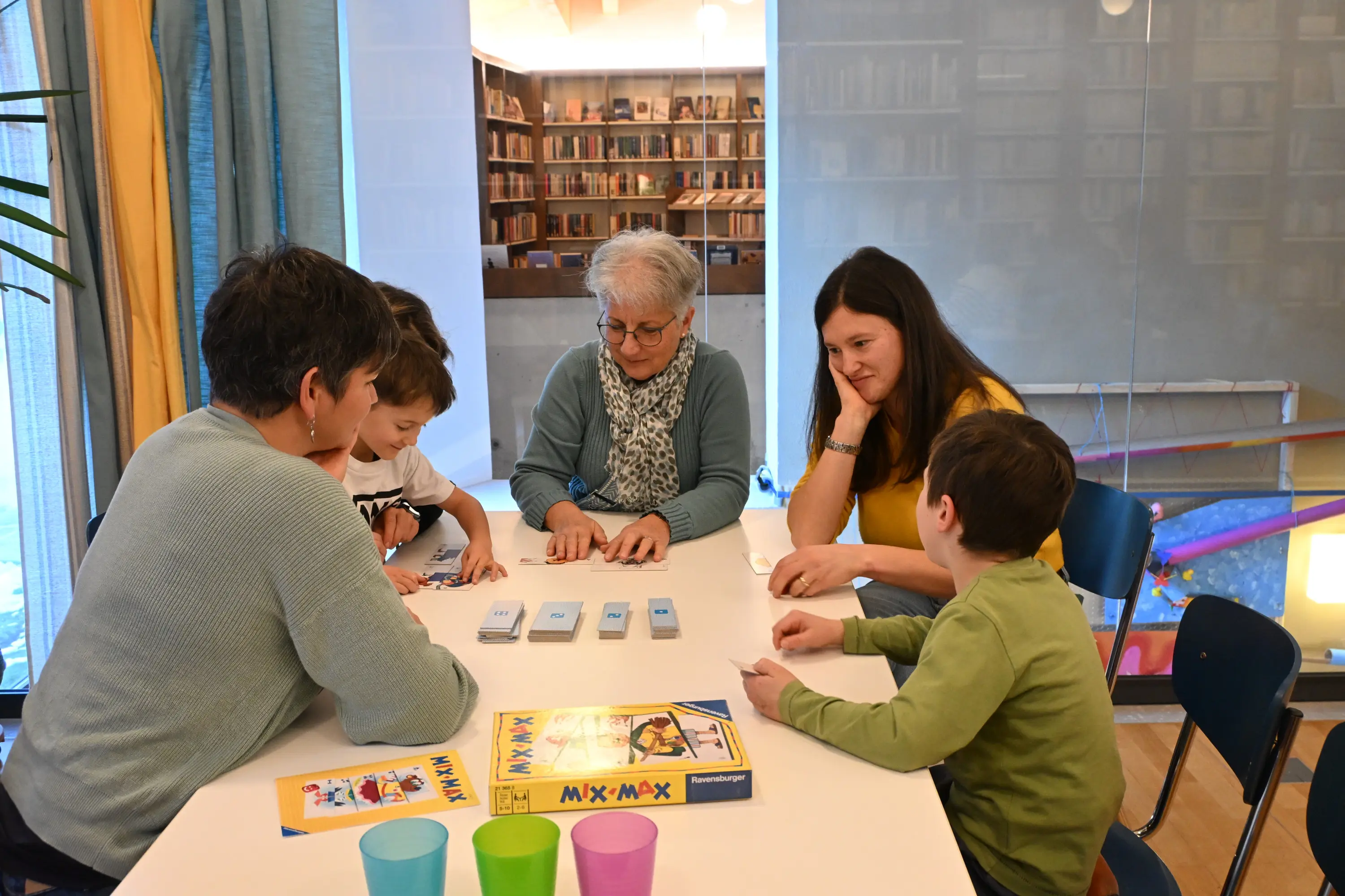 Un gruppo di quattro adulti e due bambini si riunisce attorno a un tavolo, concentrati su un gioco da tavolo. Ci sono carte sparse sul tavolo e un gioco chiam