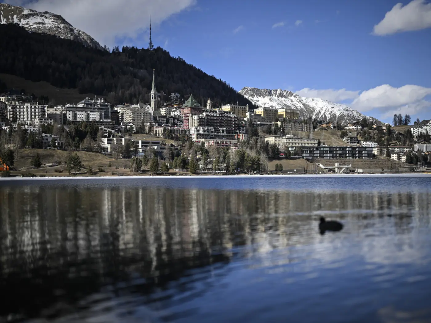 Panorama di un lago con montagne innevate sullo sfondo. Una cittadina con edifici colorati si affaccia sul lago, riflettendosi sull'acqua calma. Un'anatra