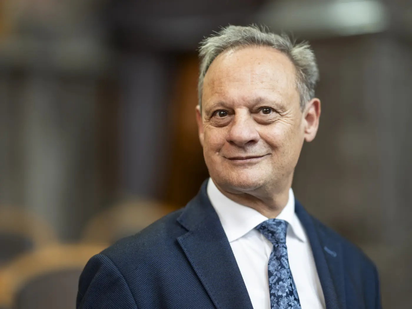 Uomo anziano con capelli grigi, indossa un abito blu scuro e una cravatta con motivo floreale. Sorridente, si trova in un ambiente elegante, con sf