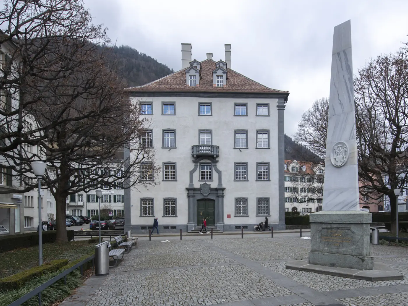 Edificio con facciata bianca e balcone in un'area urbana, circondato da alberi senza foglie. A sinistra, monumento in pietra e pavimentazione