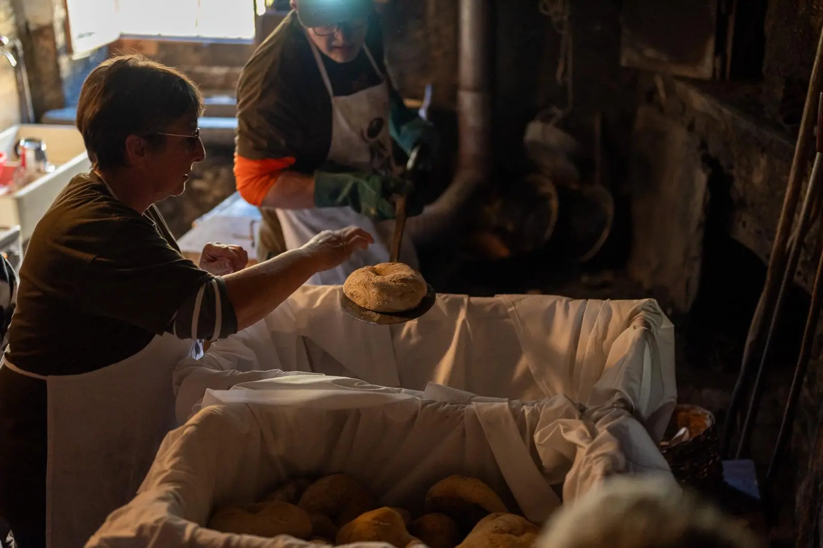 Due persone sono intente a lavorare in un ambiente rustico, mentre una solleva un pane appena sfornato sopra una cesta bianca piena di altri pani. La luce è cal