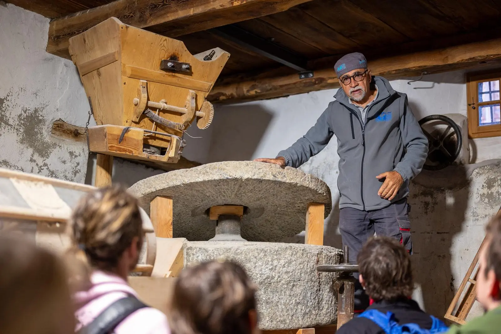 Un uomo spiega il funzionamento di un mulino tradizionale mentre un gruppo di persone ascolta attentamente. Sullo sfondo si vede un mulino in legno con una grande ru