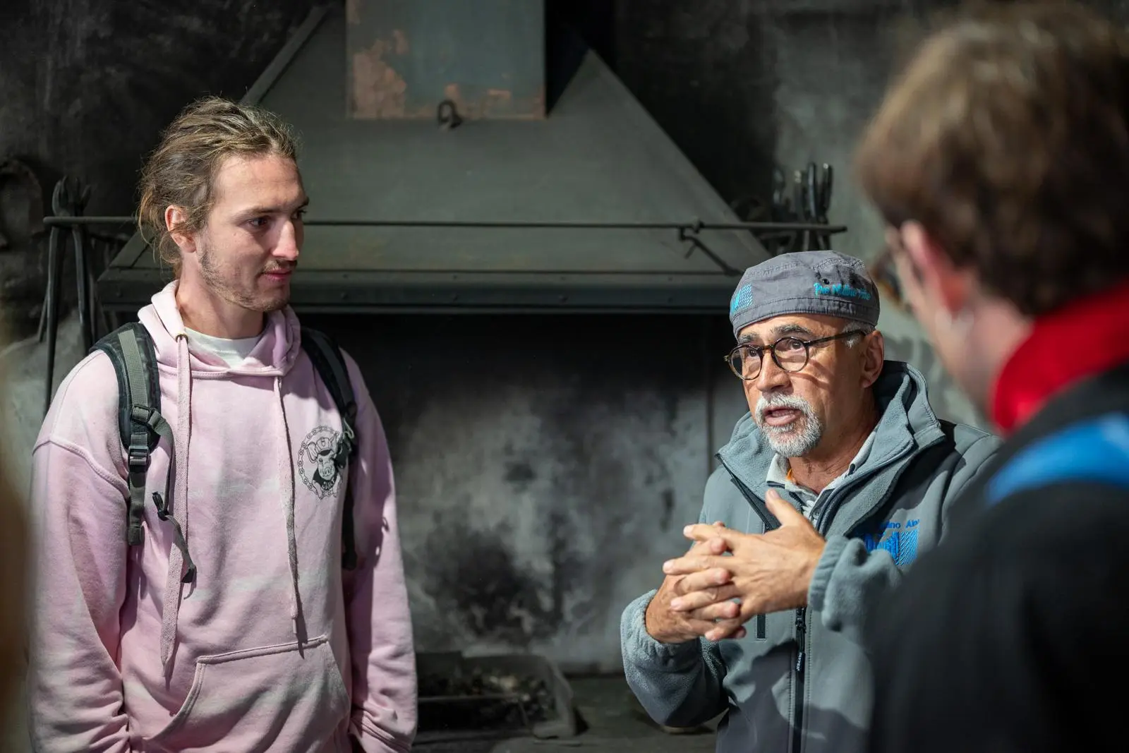Due uomini stanno conversando in un ambiente scuro e rustico, con un camino sullo sfondo. Uno dei due indossa un berretto e parla con gesti espressivi, mentre