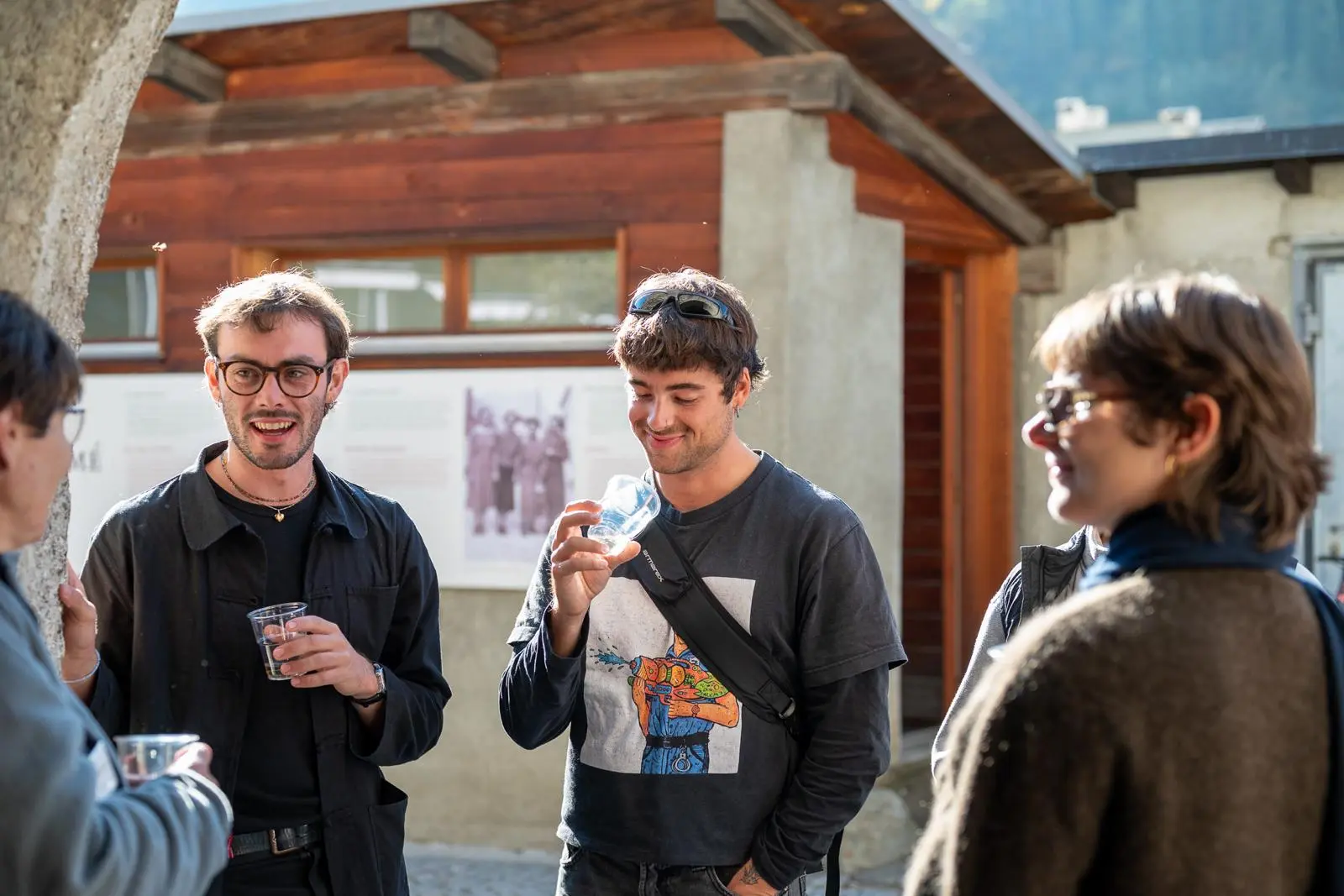 Gruppo di giovani che chiacchierano e ridono all'aperto, mentre alcuni tengono bicchieri in mano. Sullo sfondo si vede un edificio in legno e fotografie